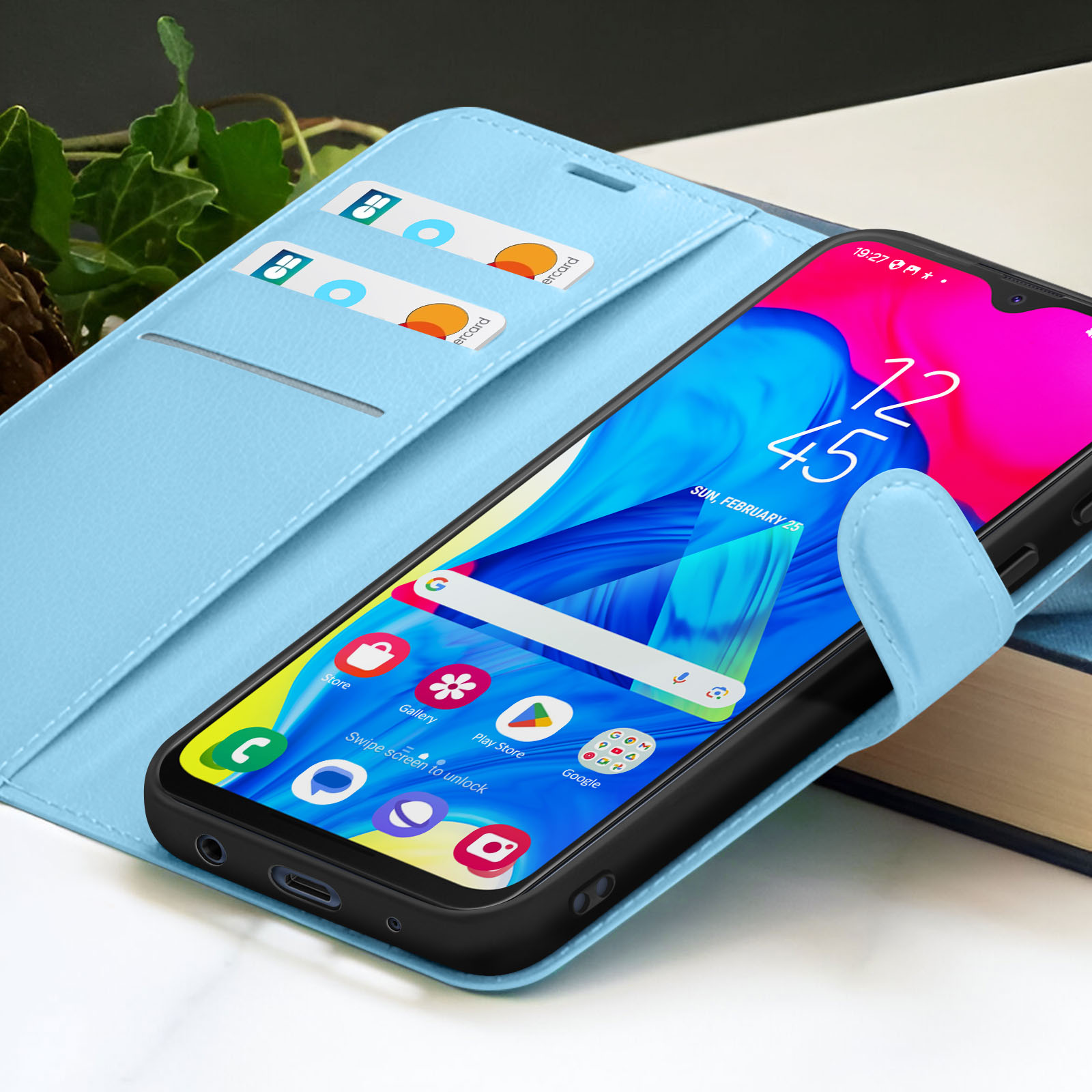Un smartphone dans un étui bleu affiche 12h45. L'étui a des emplacements pour cartes. Arrière-plan avec livre et feuillage.