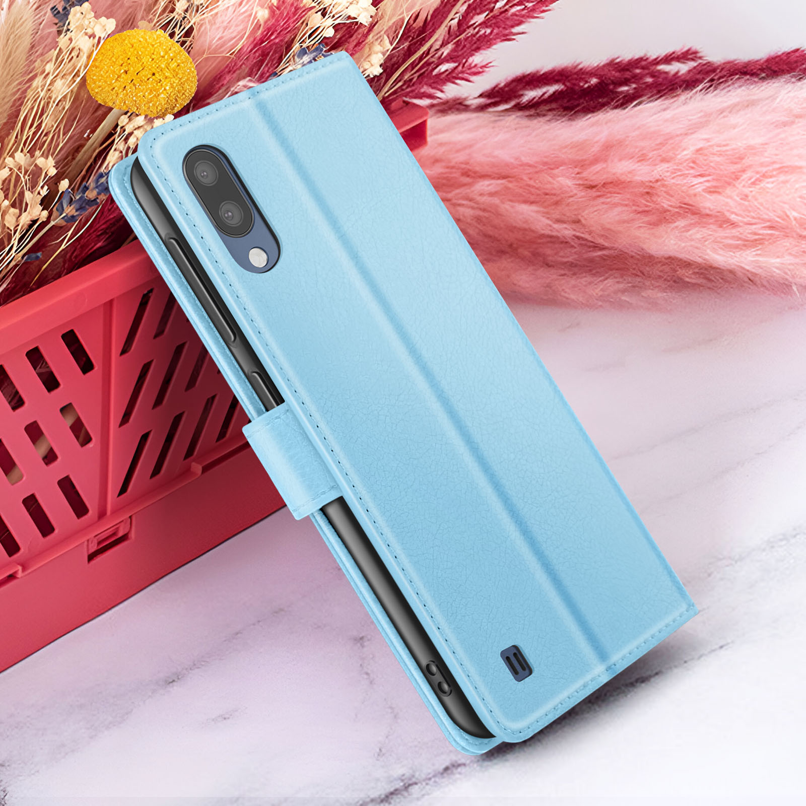Un étui de téléphone bleu clair est présenté ouvert, avec un téléphone à l'intérieur. Le fond comprend des fleurs séchées roses et rouges et un panier rouge.