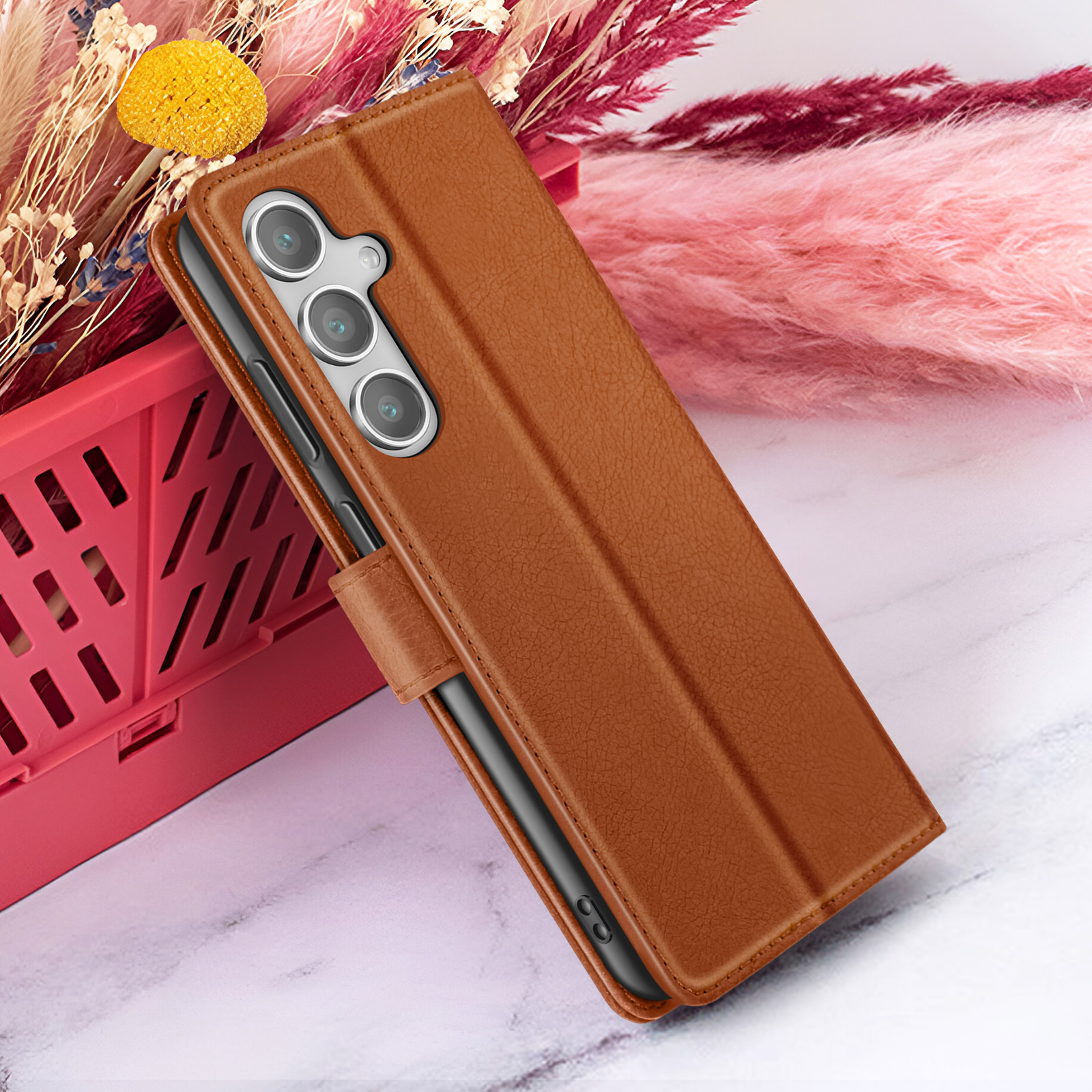 Bruine telefoonhoes met telefoon erin, rode mand, roze bloemen en gele bloem op wit oppervlak.