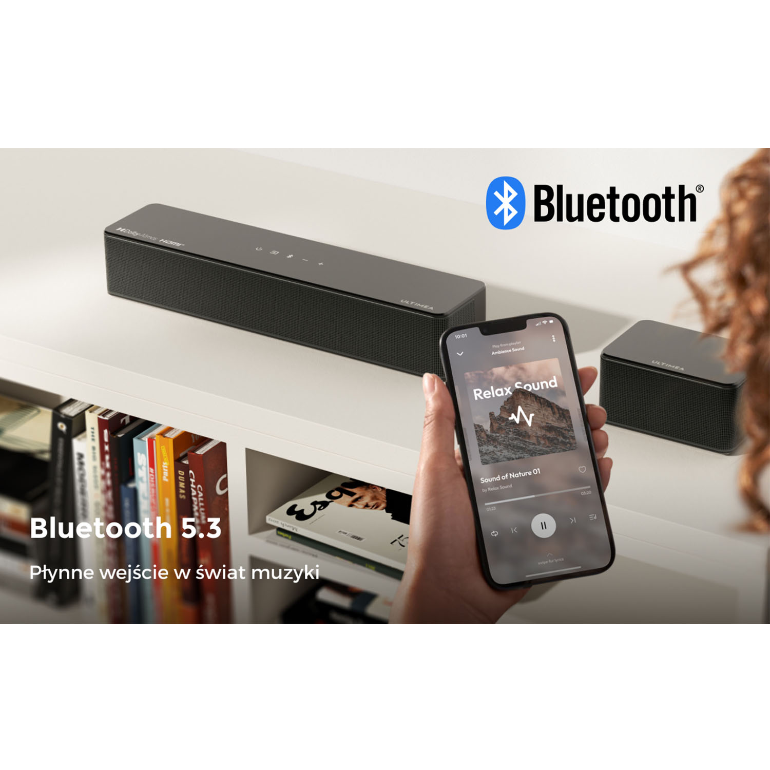 Osoba trzymająca telefon odtwarzający muzykę. Głośnik Bluetooth, książki i mały głośnik na białym stole.