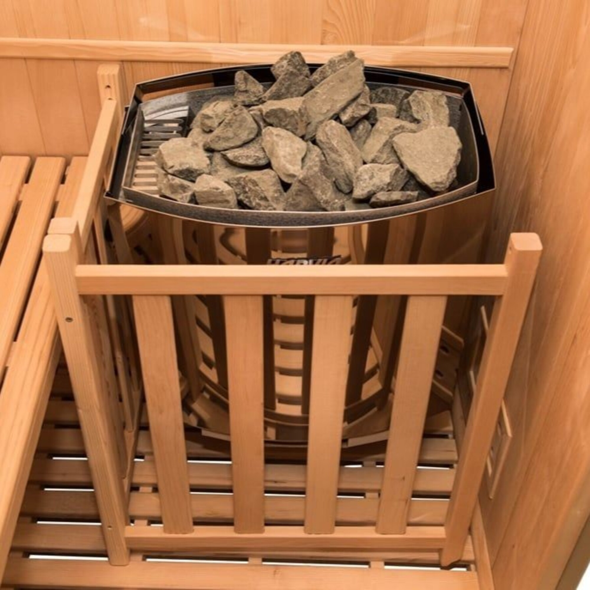 Una estufa de sauna con rocas dentro. Está hecha de acero inoxidable y rodeada de soportes y listones de madera.
