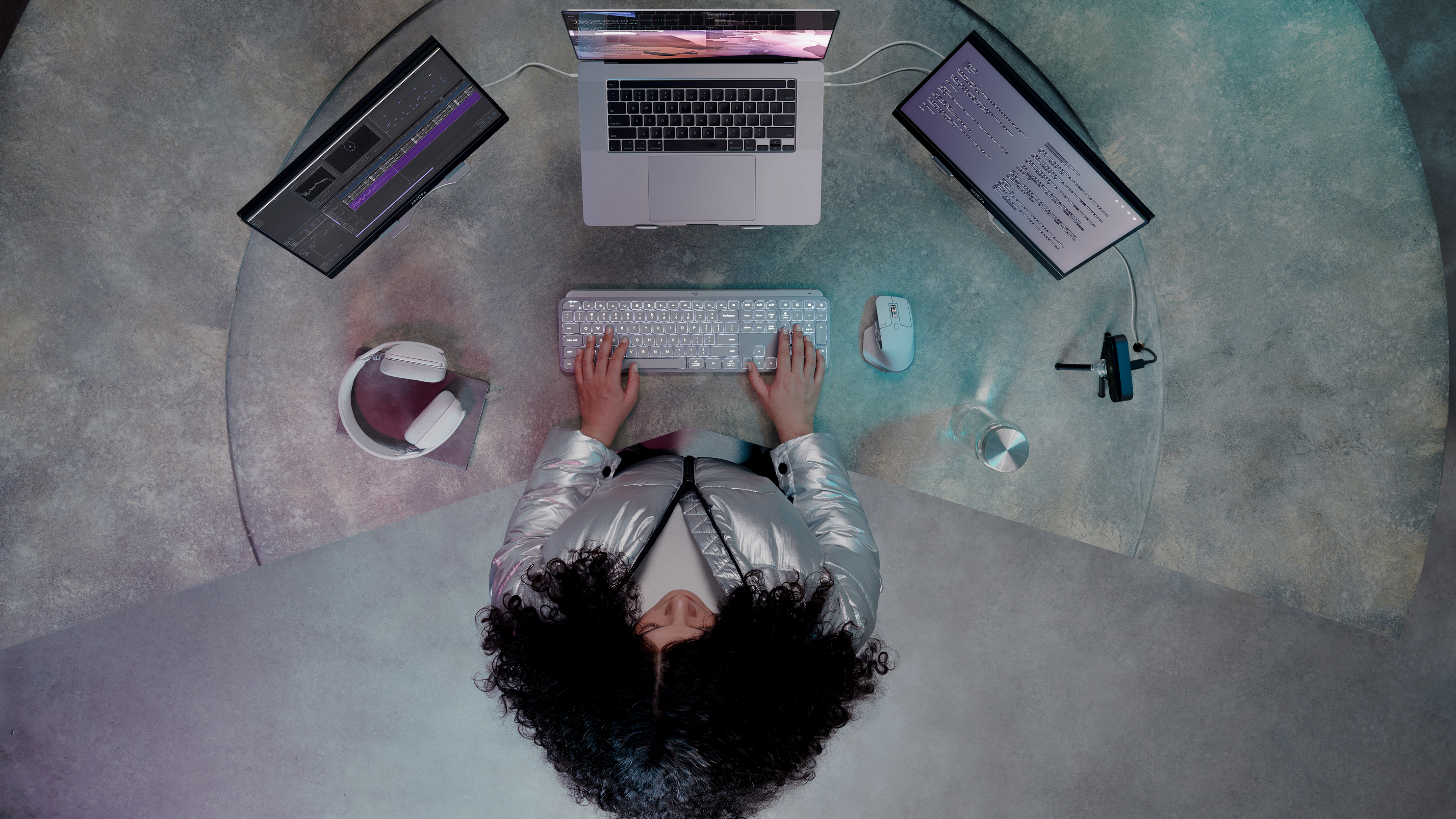 Widok z góry na osobę przy biurku z wieloma ekranami, laptopem, klawiaturą i słuchawkami. Osoba ma na sobie srebrną kurtkę.