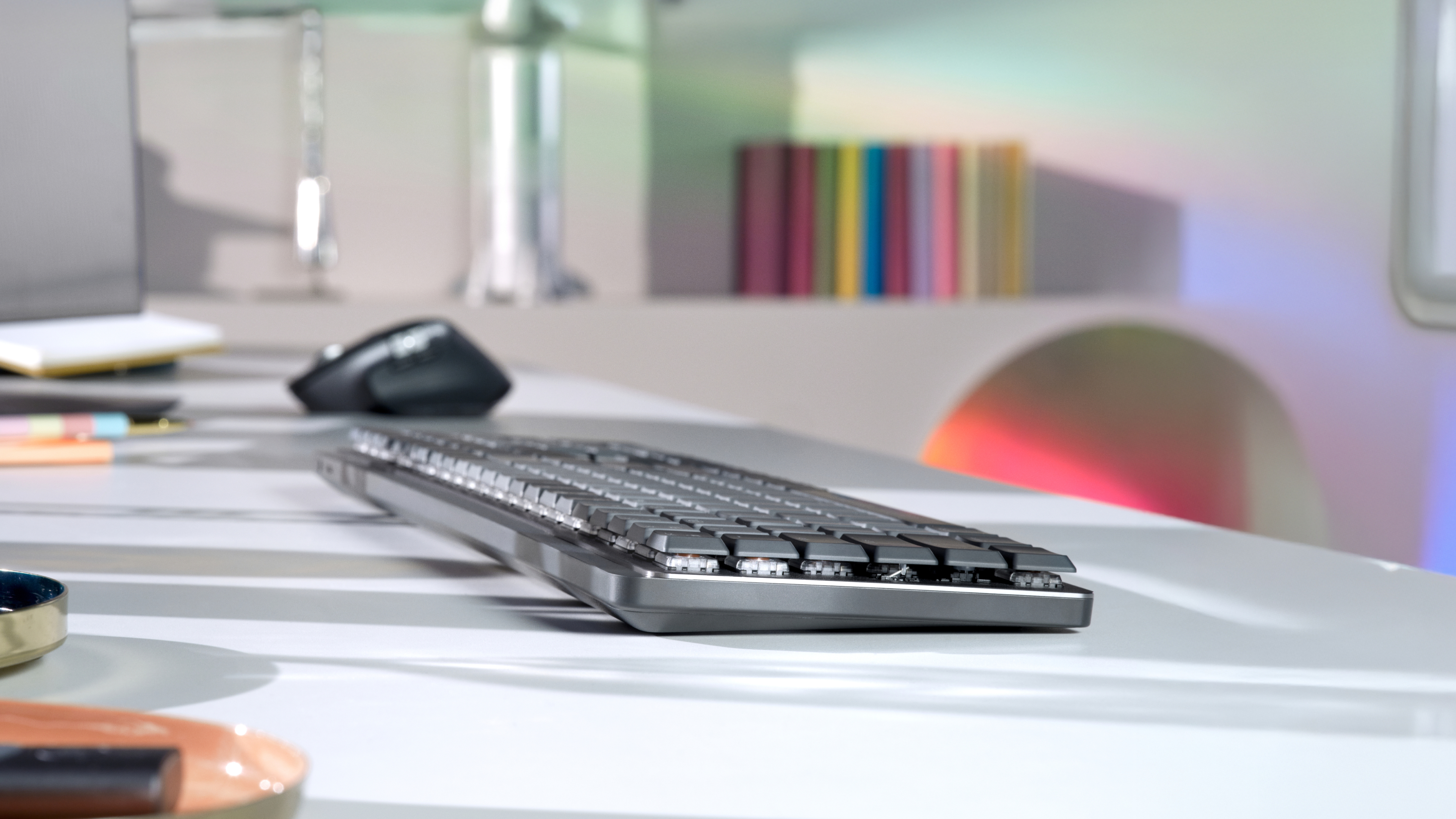 Szara klawiatura na białym biurku. Widoczna mysz i laptop. Książki w tle z kolorowymi grzbietami.