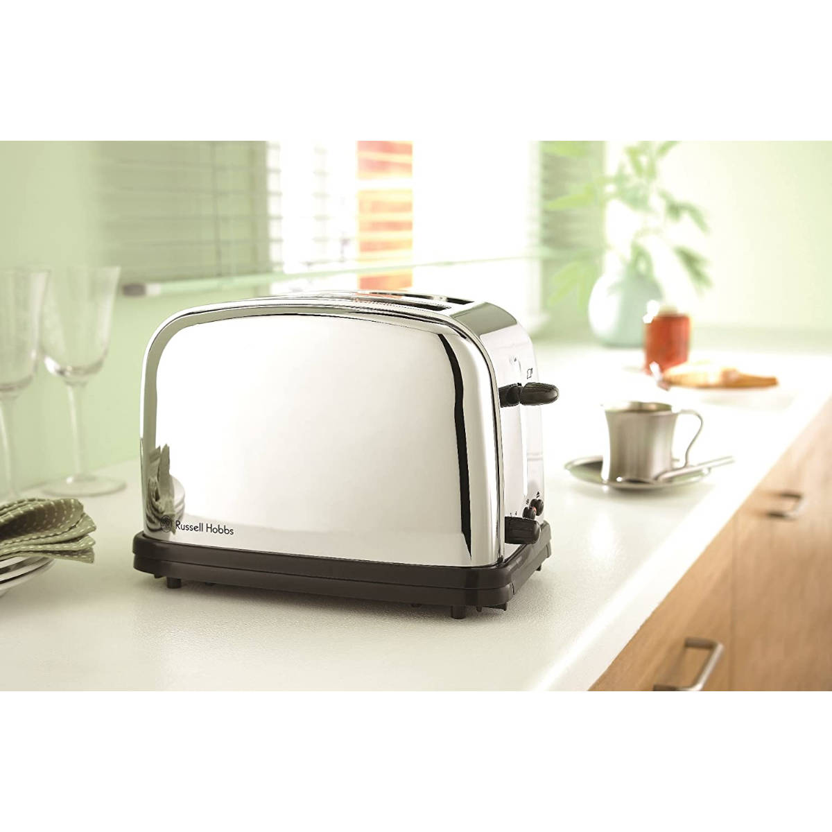 Ein verchromter Toaster mit schwarzem Sockel steht auf einer weißen Theke. Eine Kaffeetasse, Weingläser und ein Marmeladenglas sind sichtbar.