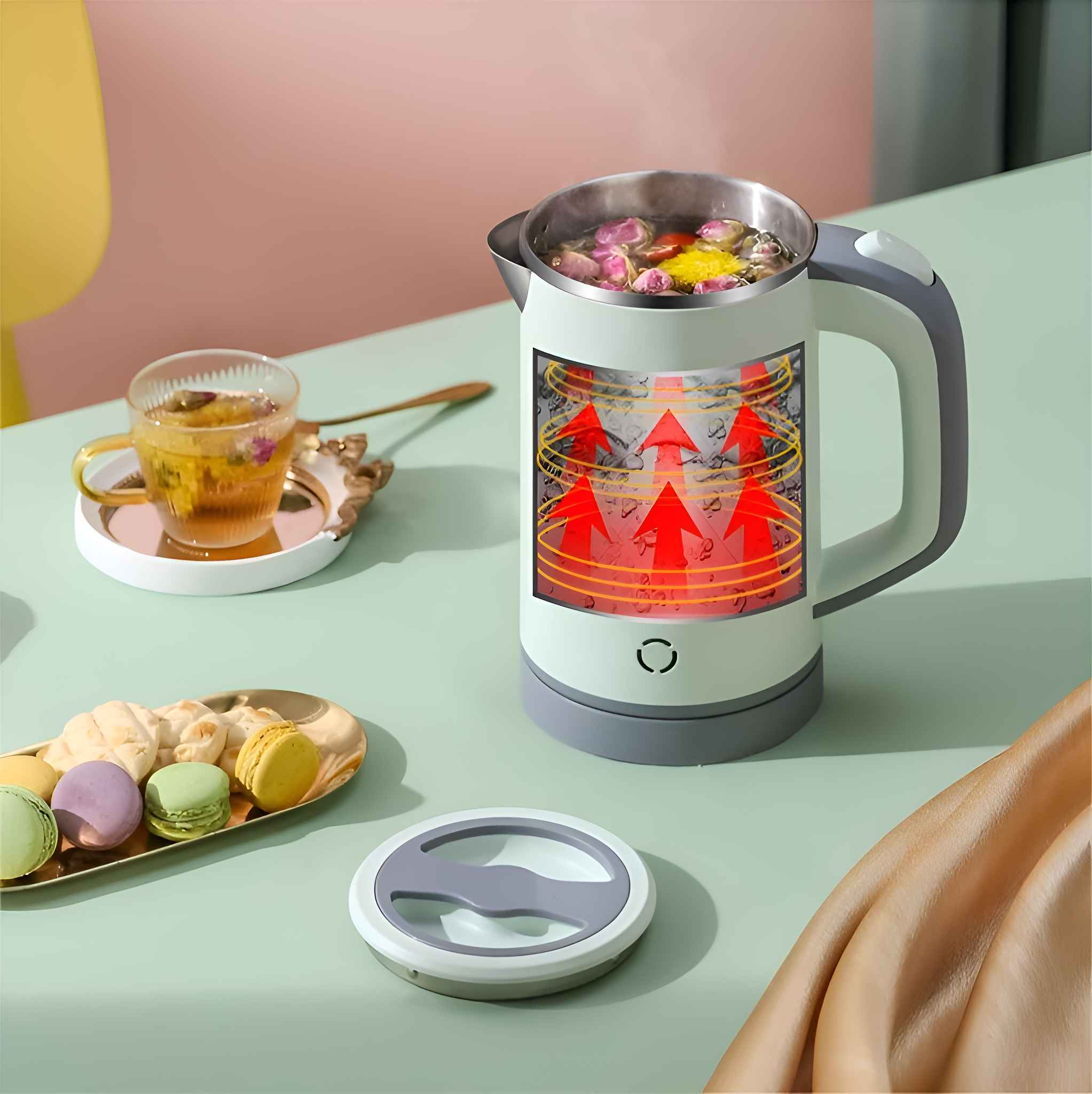 Ein weißer Wasserkocher mit einer Tasse Tee, Makronen und Blumen im Inneren auf einem grünen Tisch.