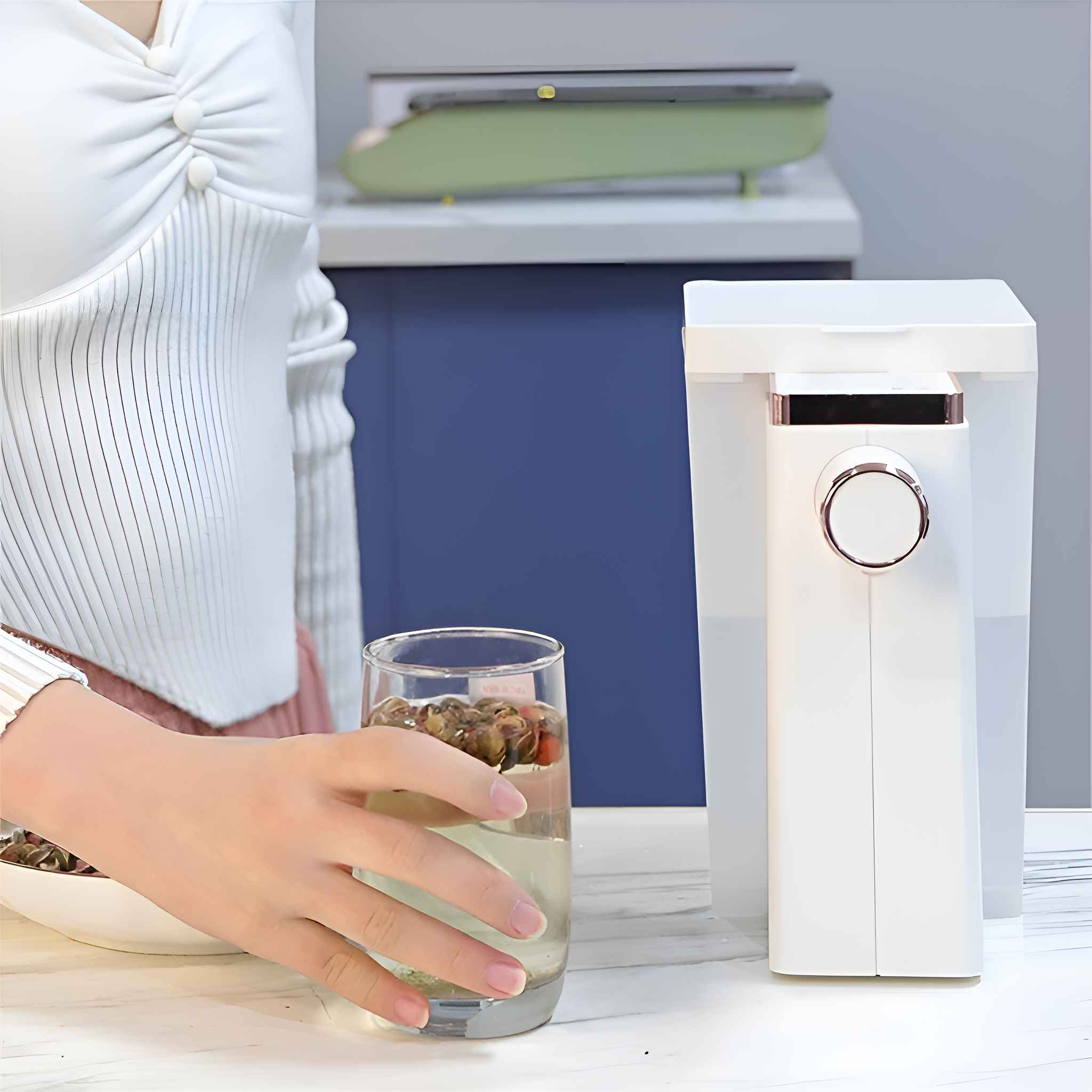 Eine Person hält ein Glas Wasser mit Trockenfrüchten. Rechts befindet sich ein weißer Wasserspender, im Hintergrund ein grünes Objekt.