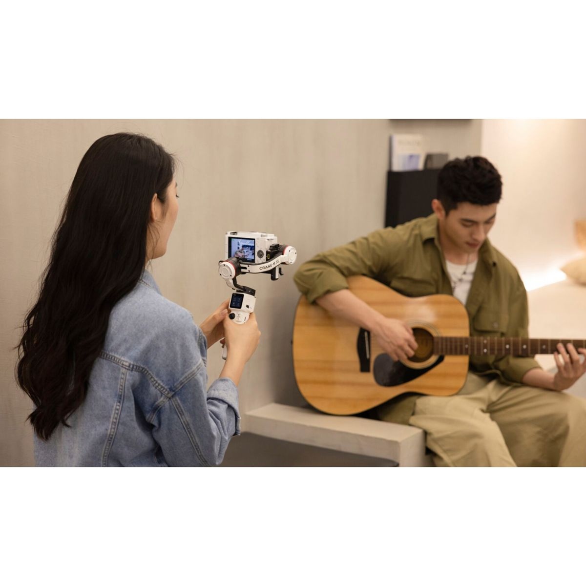Eine Frau filmt einen Mann, der Gitarre spielt. Sie hält eine Kamera auf einem Gimbal. Mann spielt Akustikgitarre, drinnen, in ungezwungener Atmosphäre.