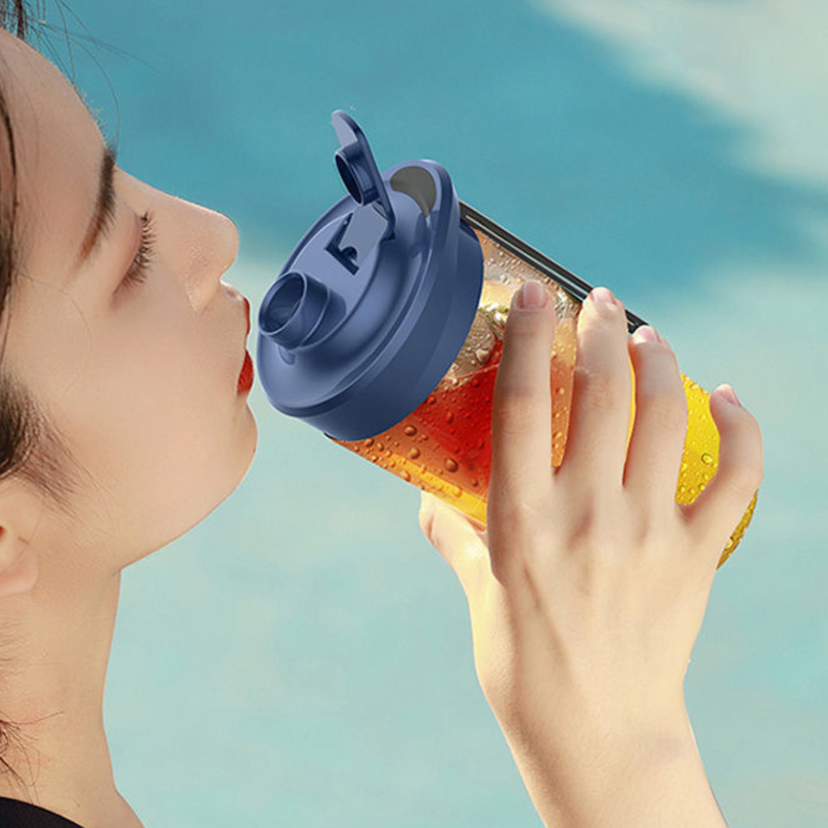 Eine Frau trinkt aus einer blau verschlossenen Flasche mit orangener Flüssigkeit. Der Hintergrund ist ein verschwommener blauer Himmel.