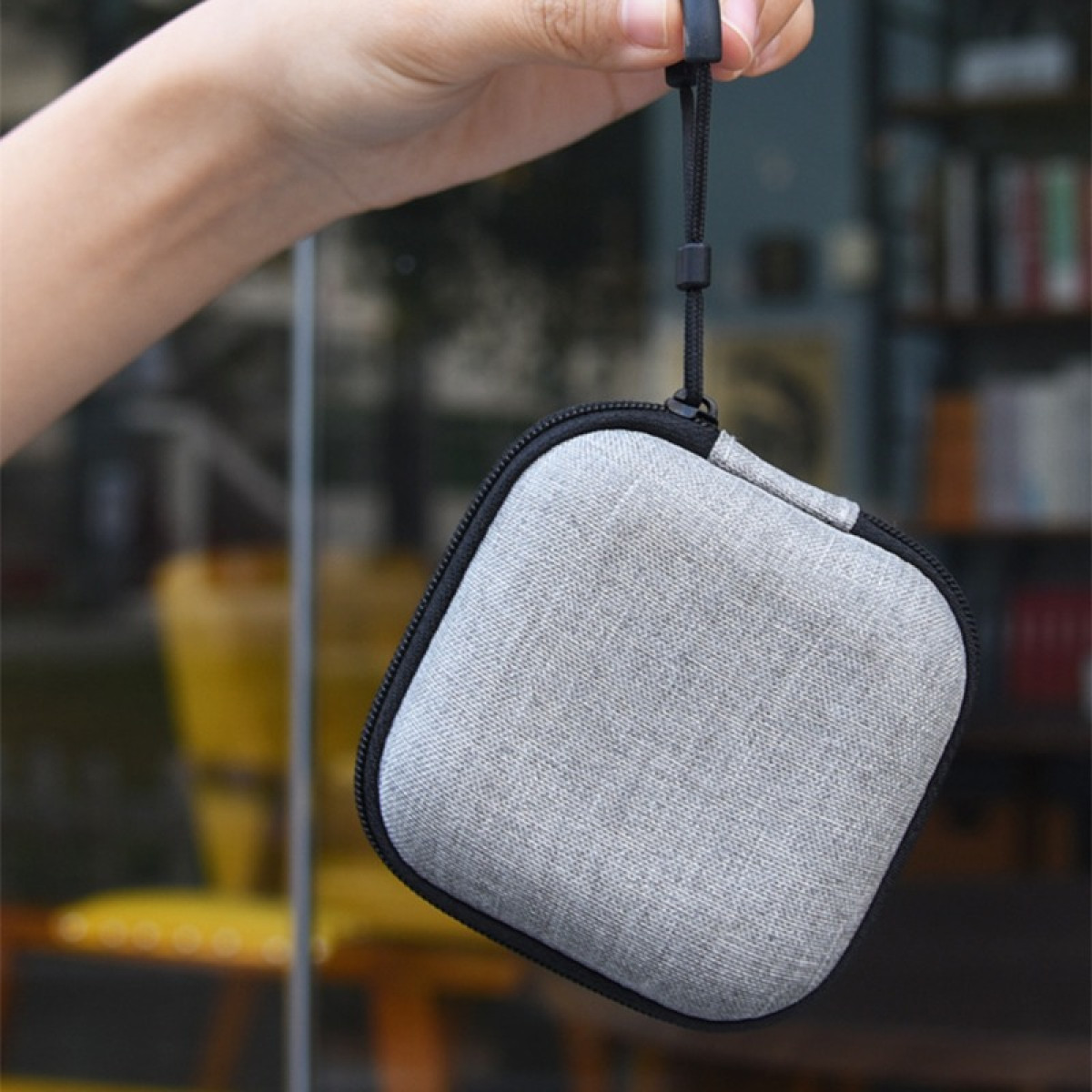 Una mano sostiene una funda gris cuadrada con cremallera y correa negras. El fondo muestra una estantería borrosa y una silla amarilla.