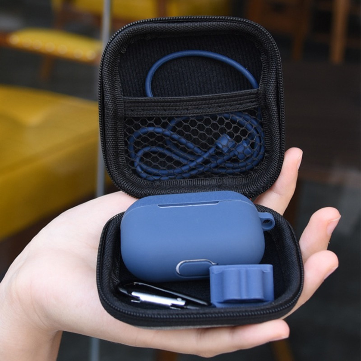 Una mano sostiene un estuche negro abierto con auriculares azules, un cable de carga y accesorios. El fondo está borroso.