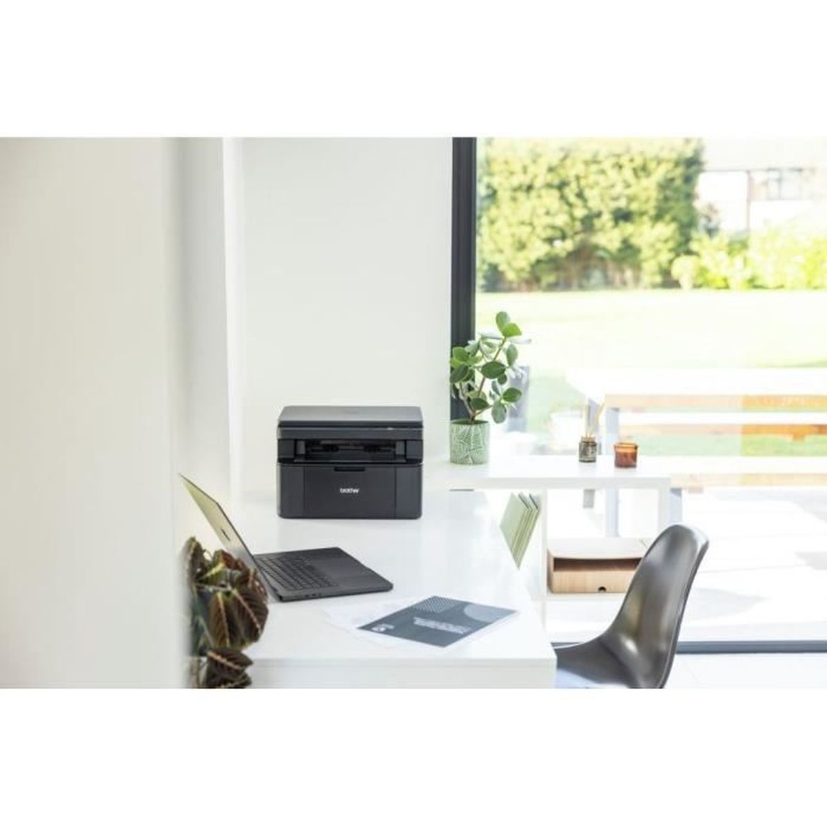 Een thuiskantoor met een zwarte printer, laptop en plant op een wit bureau. Een raam toont de tuin.