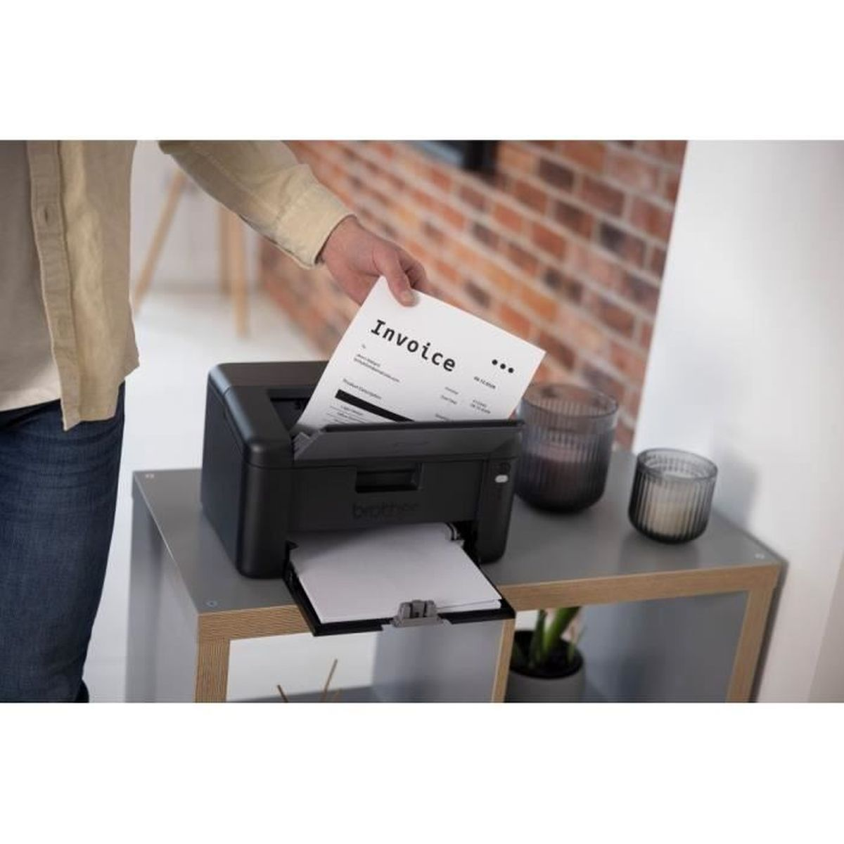 Osoba wkłada fakturę do czarnej drukarki. Drukarka stoi na szarym stole ze świecami i rośliną. W tle ceglana ściana.