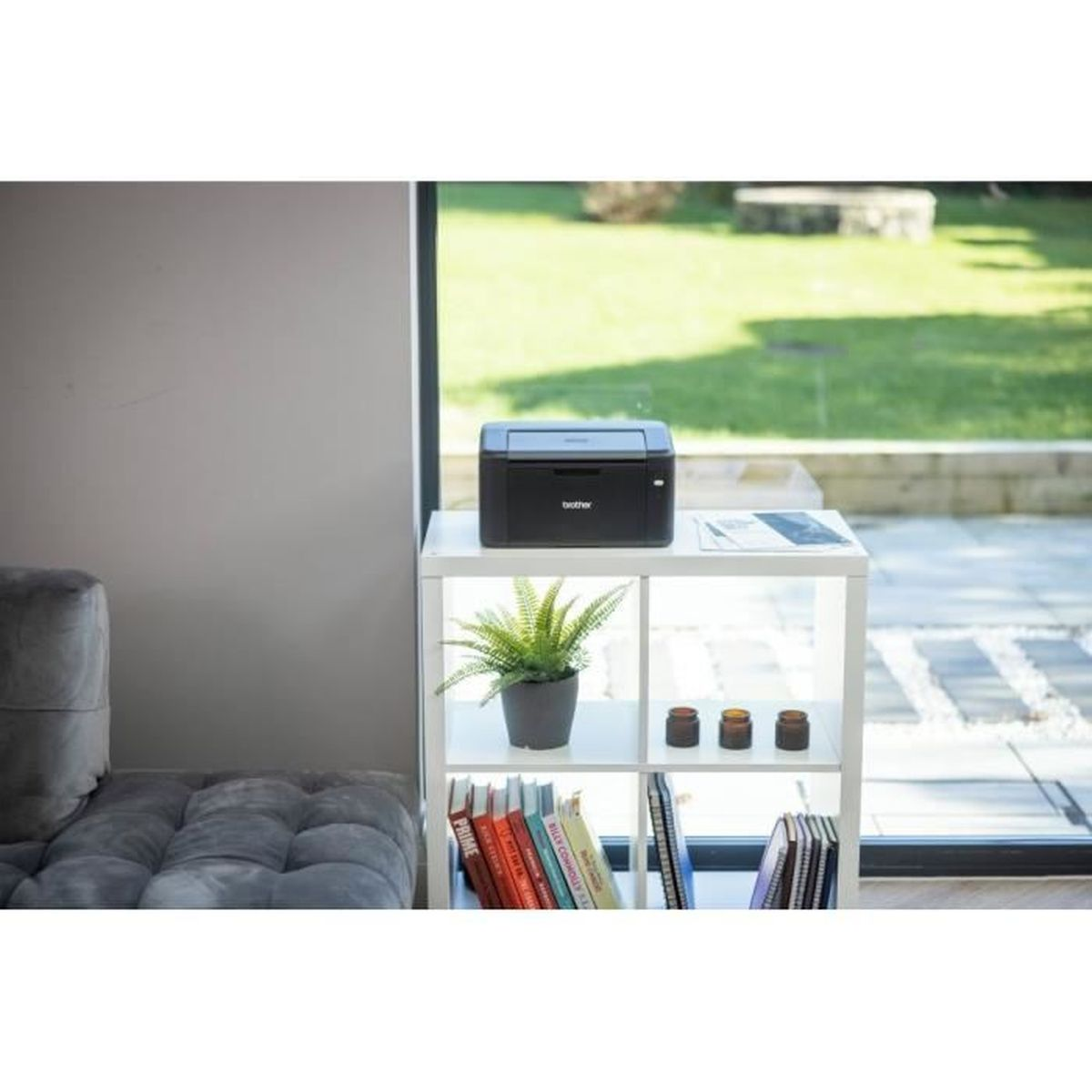 Czarna drukarka na białej półce z doniczką, książkami i trzema małymi słoiczkami. Okno pokazuje zielony trawnik.