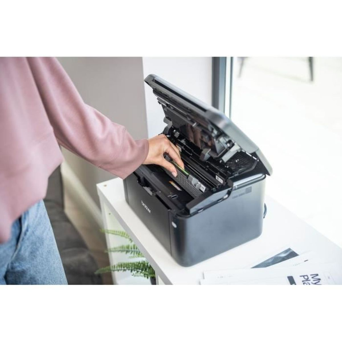Osoba wkłada wkład z tonerem do czarnej drukarki. Drukarka stoi na białej półce. W tle widać okno.