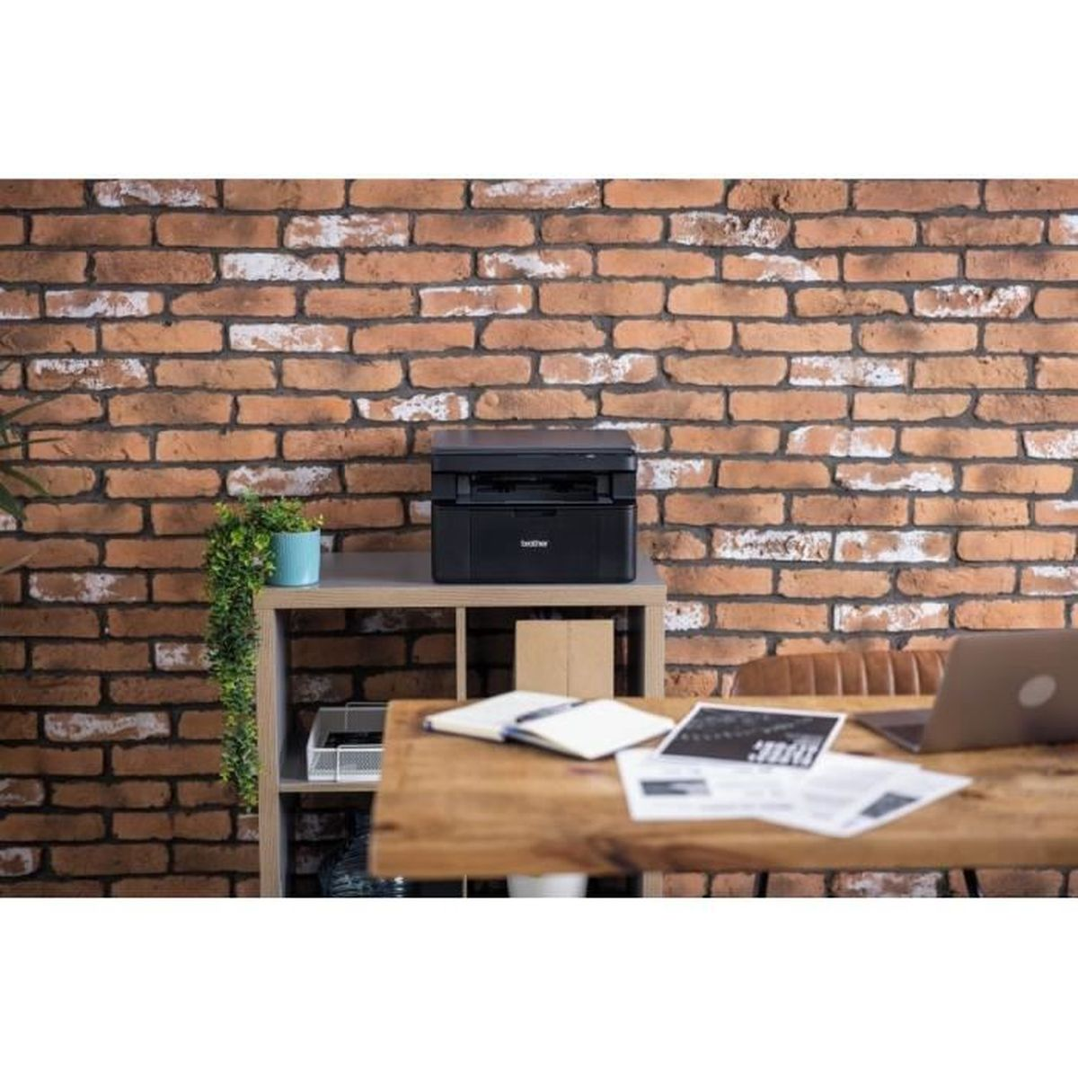 Czarna drukarka Brother stoi na półce. Przed nią biurko z laptopem i papierami. Ceglana ściana stanowi tło.