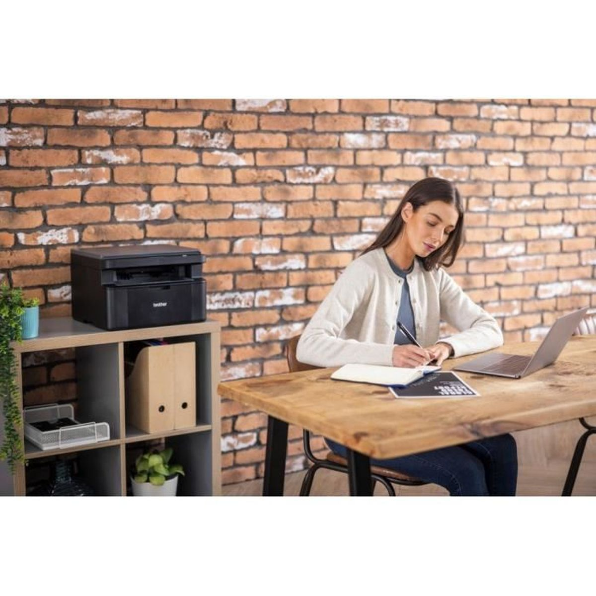 Kobieta przy biurku z laptopem i notatnikiem, pisząca. Drukarka i półka po lewej. Tło z ceglanego muru.