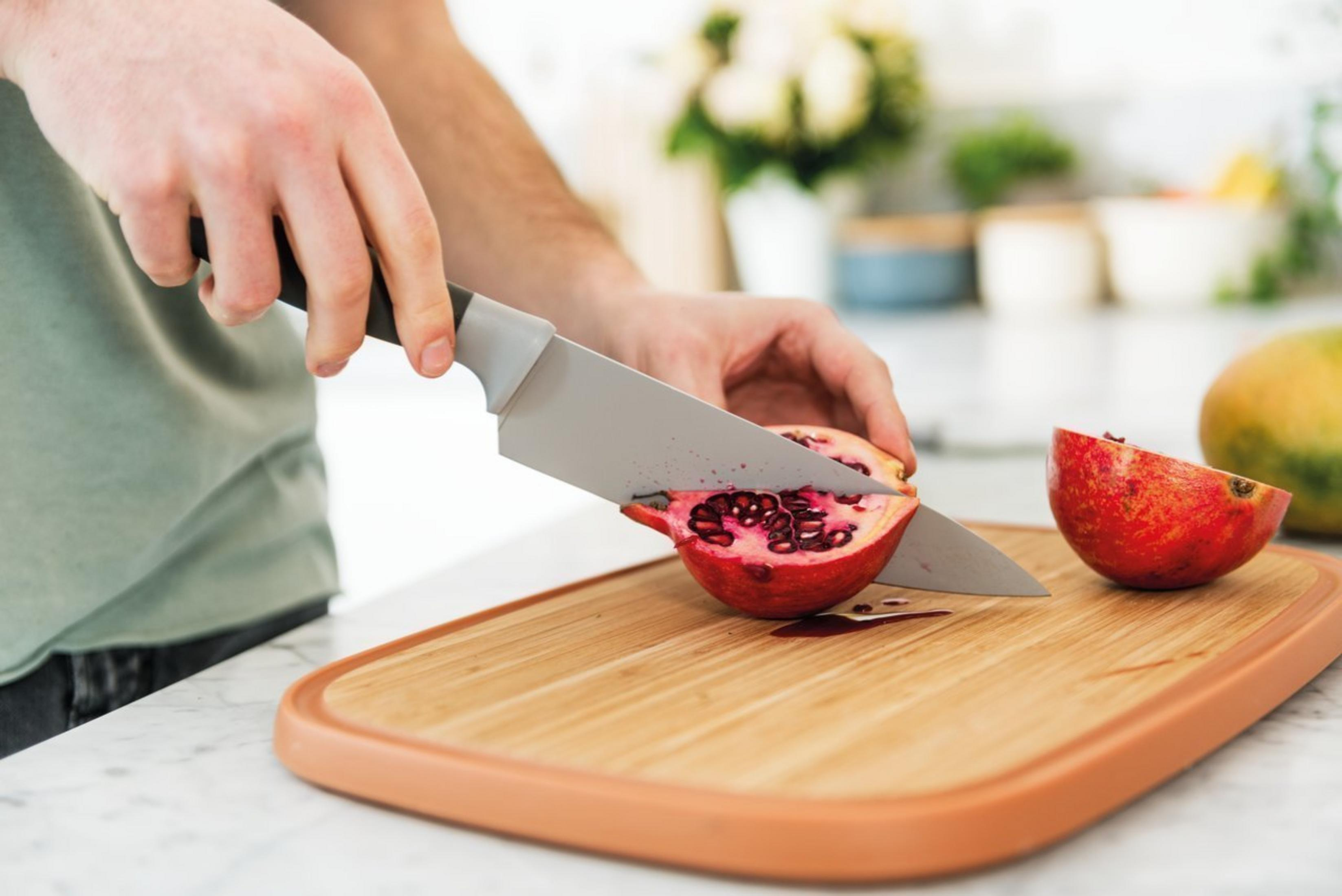 A person cuts a pomegranate on a wooden board with a knife. A cut pomegranate piece lies beside.