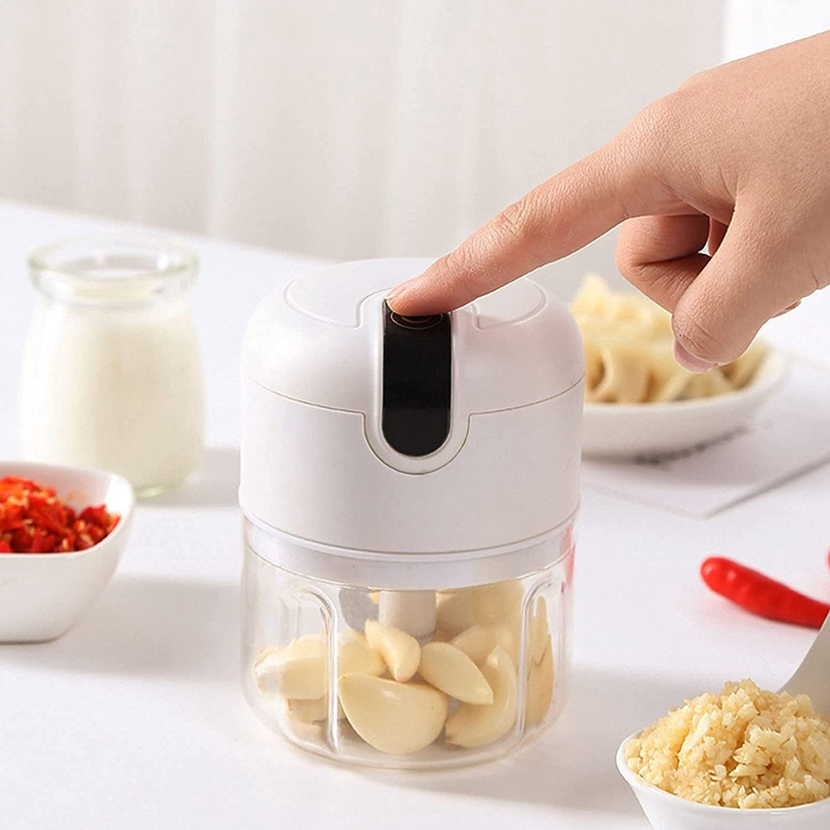 A hand presses a button on a white garlic chopper. Chopped garlic, red peppers, and a glass of milk are in the background.