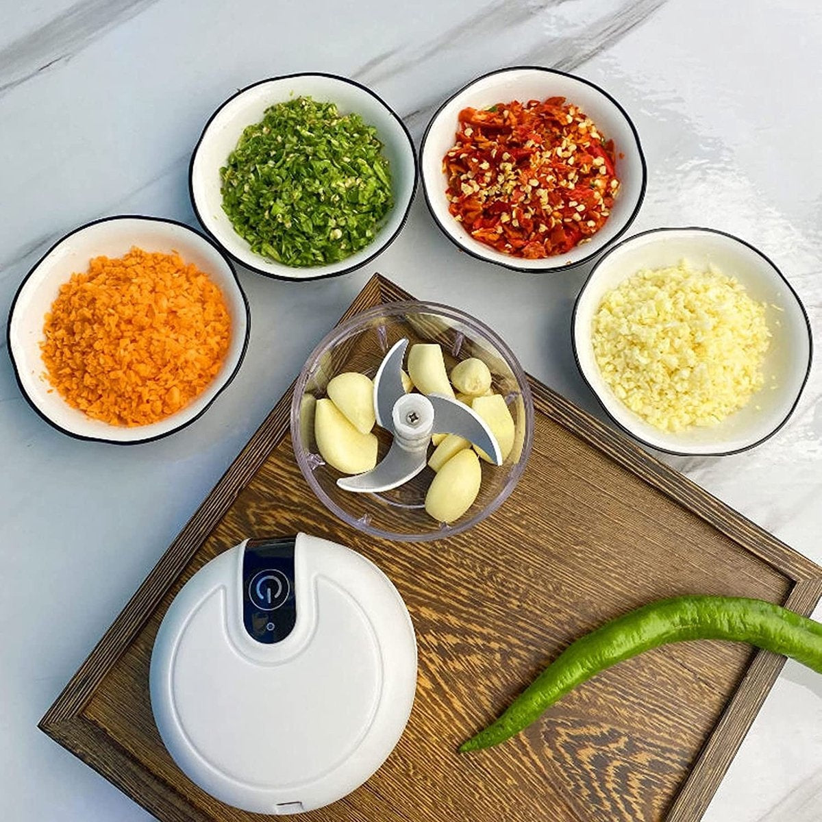 Chopped vegetables in bowls, garlic in a chopper, and a green pepper on a wooden tray. A white chopper is also present.