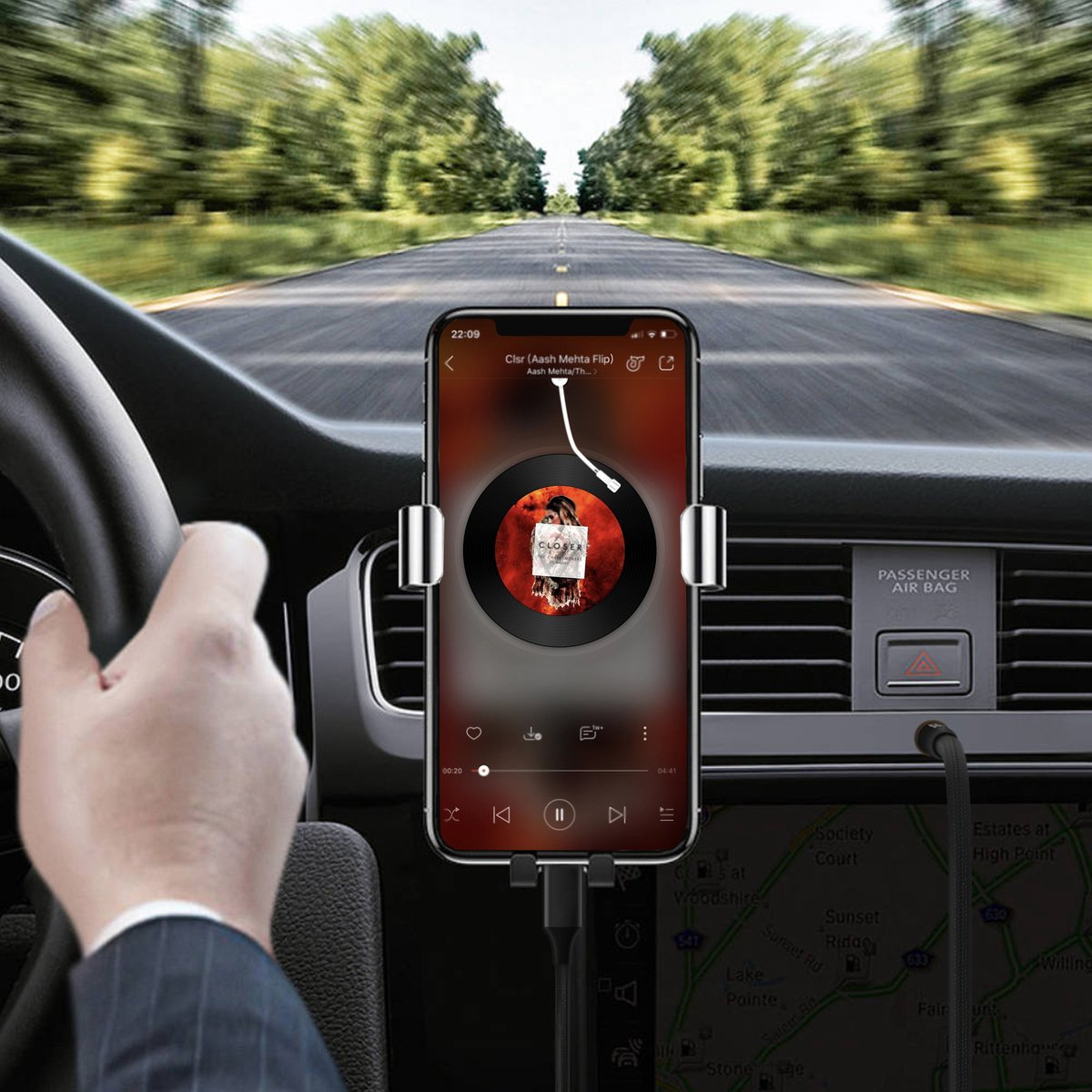 Phone in car mount playing music, driver's hand on steering wheel, road ahead. Trees line the road. Map visible below.
