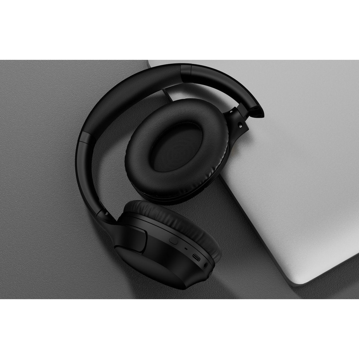 Black headphones on a gray surface, next to a laptop. The headphones are folded. The background is a light gray.