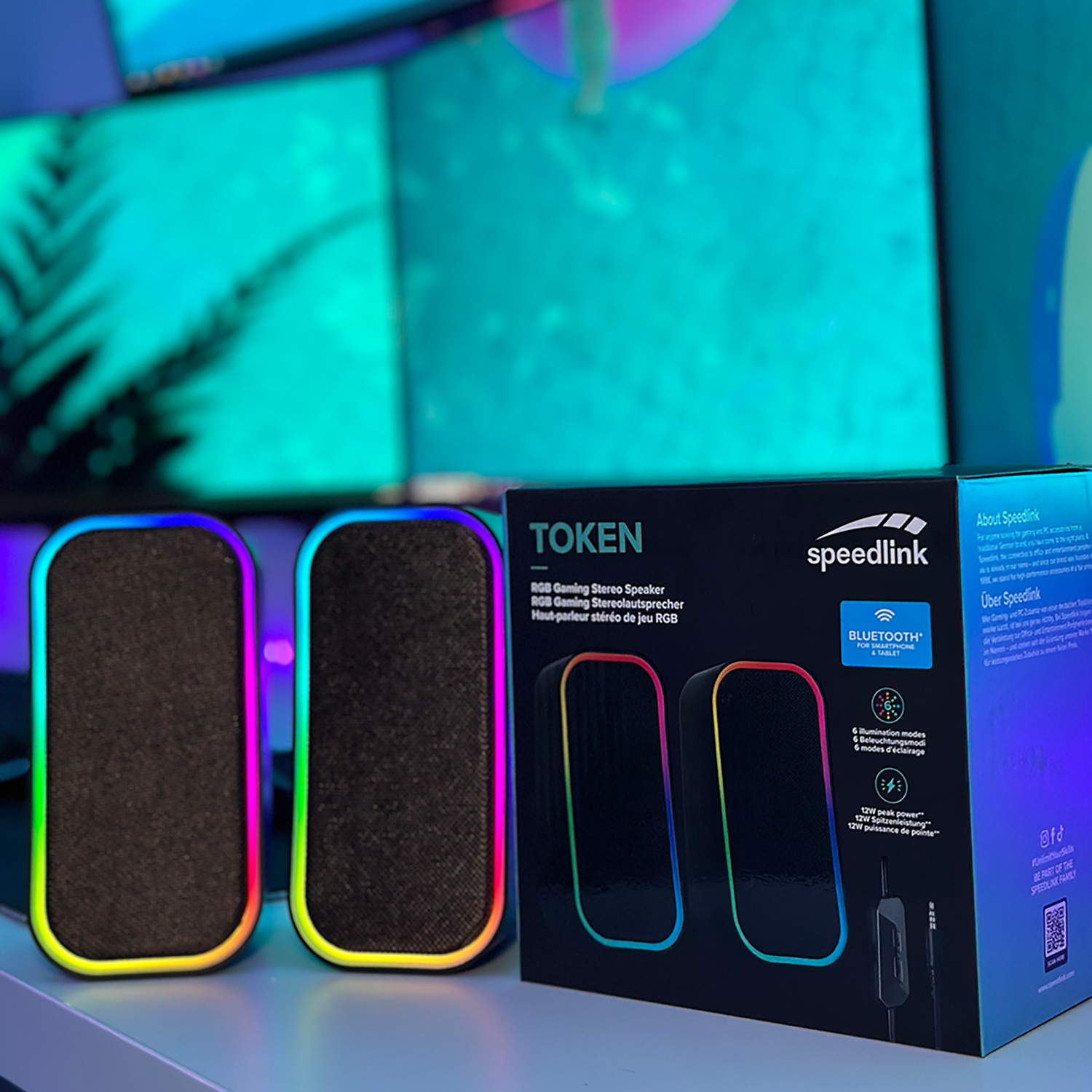 Due altoparlanti da gioco RGB e una scatola. La scatola ha l'immagine degli altoparlanti. Lo sfondo ha due monitor.