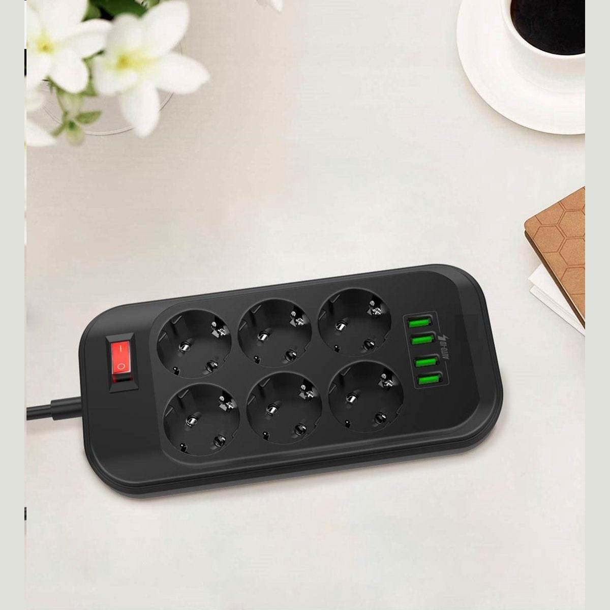 Zwarte stekkerdoos met zes stopcontacten en USB-poorten. Een rode schakelaar staat aan. Bloemen, koffie en een notitieboek op de achtergrond.