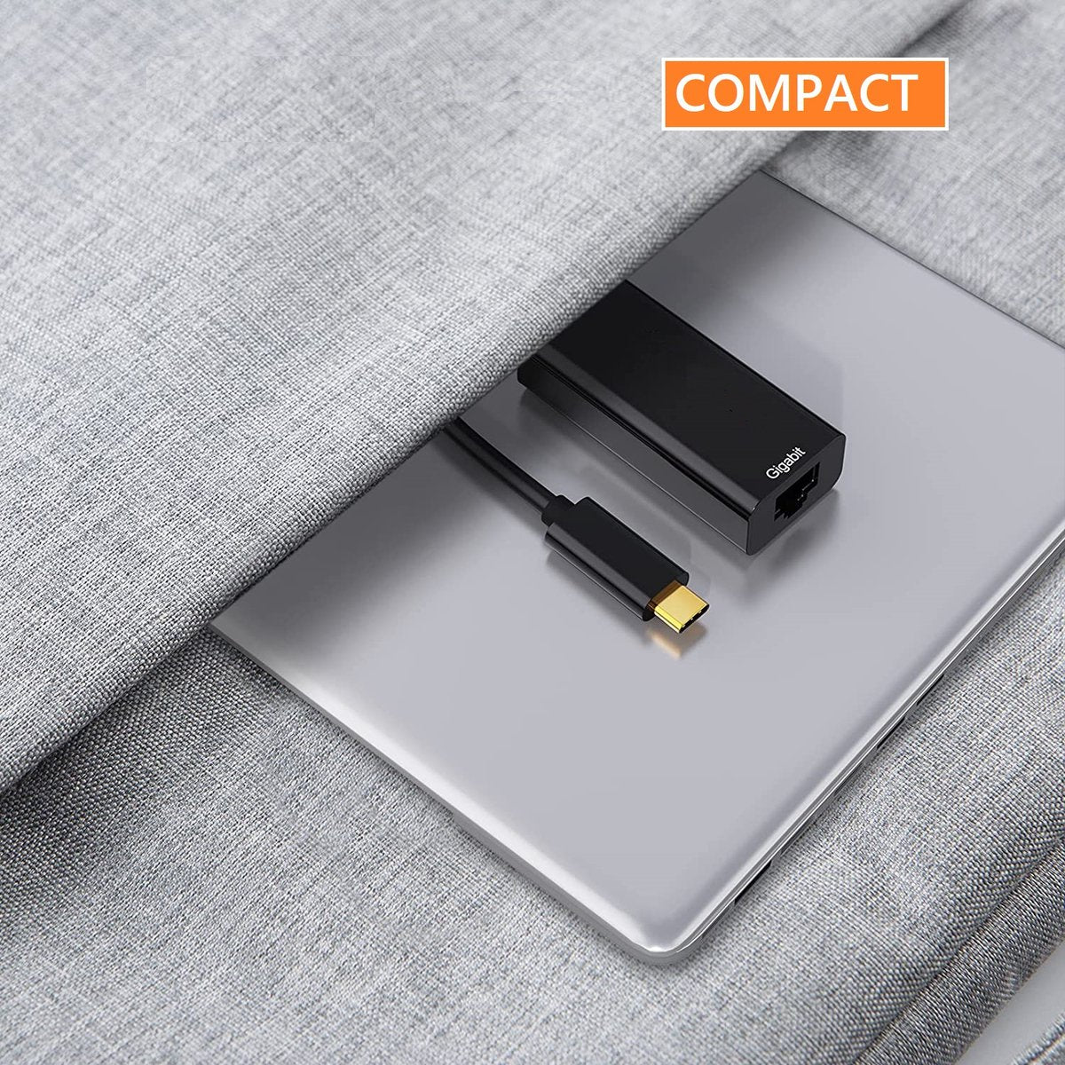 A black Gigabit ethernet adapter with a USB-C cable and a laptop on a gray surface. The word compact is on the top.