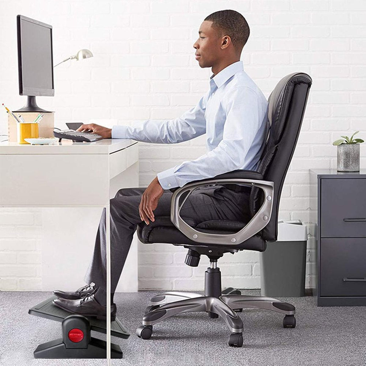 A man sits at a desk, working on a computer. He is using a footrest. The office has a white brick wall and a black chair.