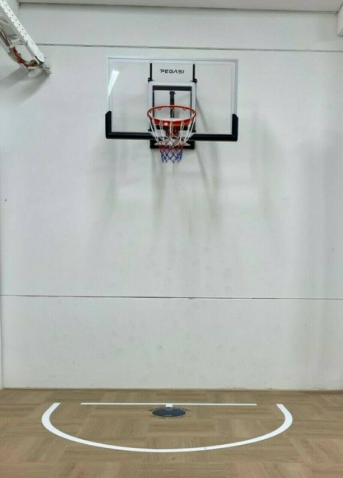 A basketball hoop with net on a white wall. The floor has a free-throw line marked. The scene is indoor.