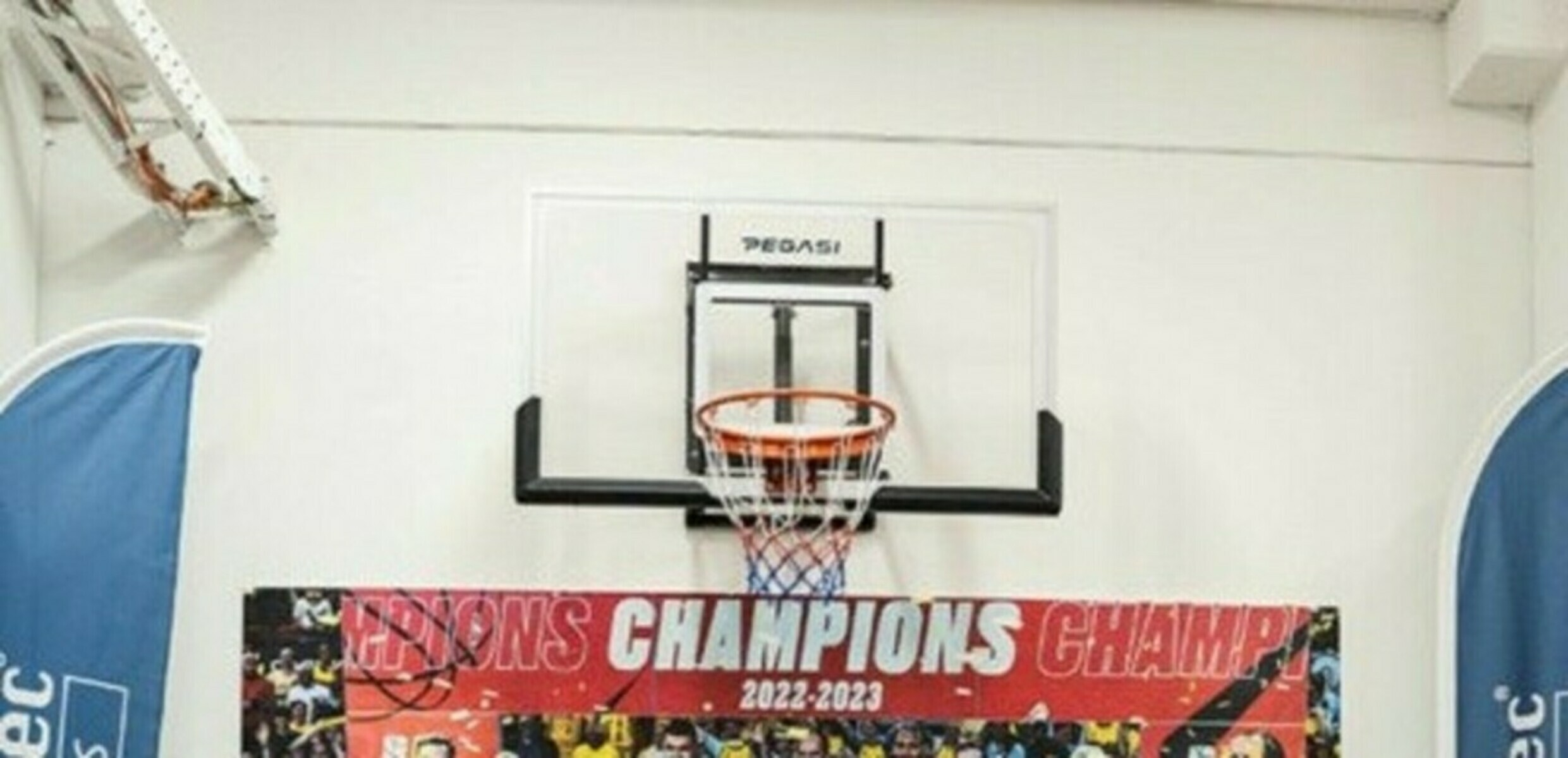 A basketball hoop is centered on a white wall. Below it is a banner that says 'Champions 2022-2023'.