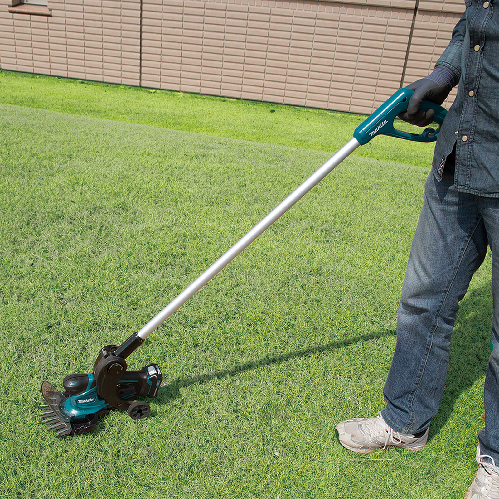 A person holds a green and silver Makita lawn edger on a green lawn. A beige wall is in the background.