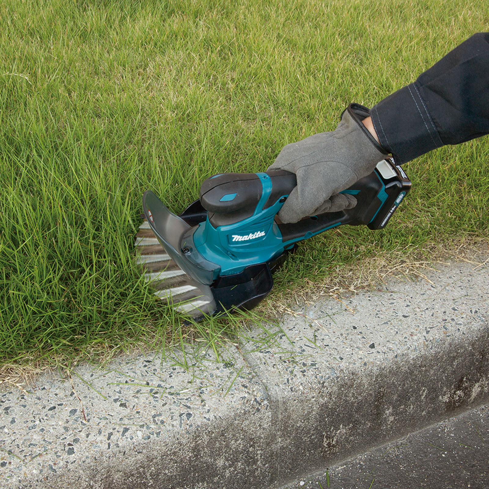 A person in a grey glove uses a Makita grass trimmer. Green grass and a concrete curb are in the scene.