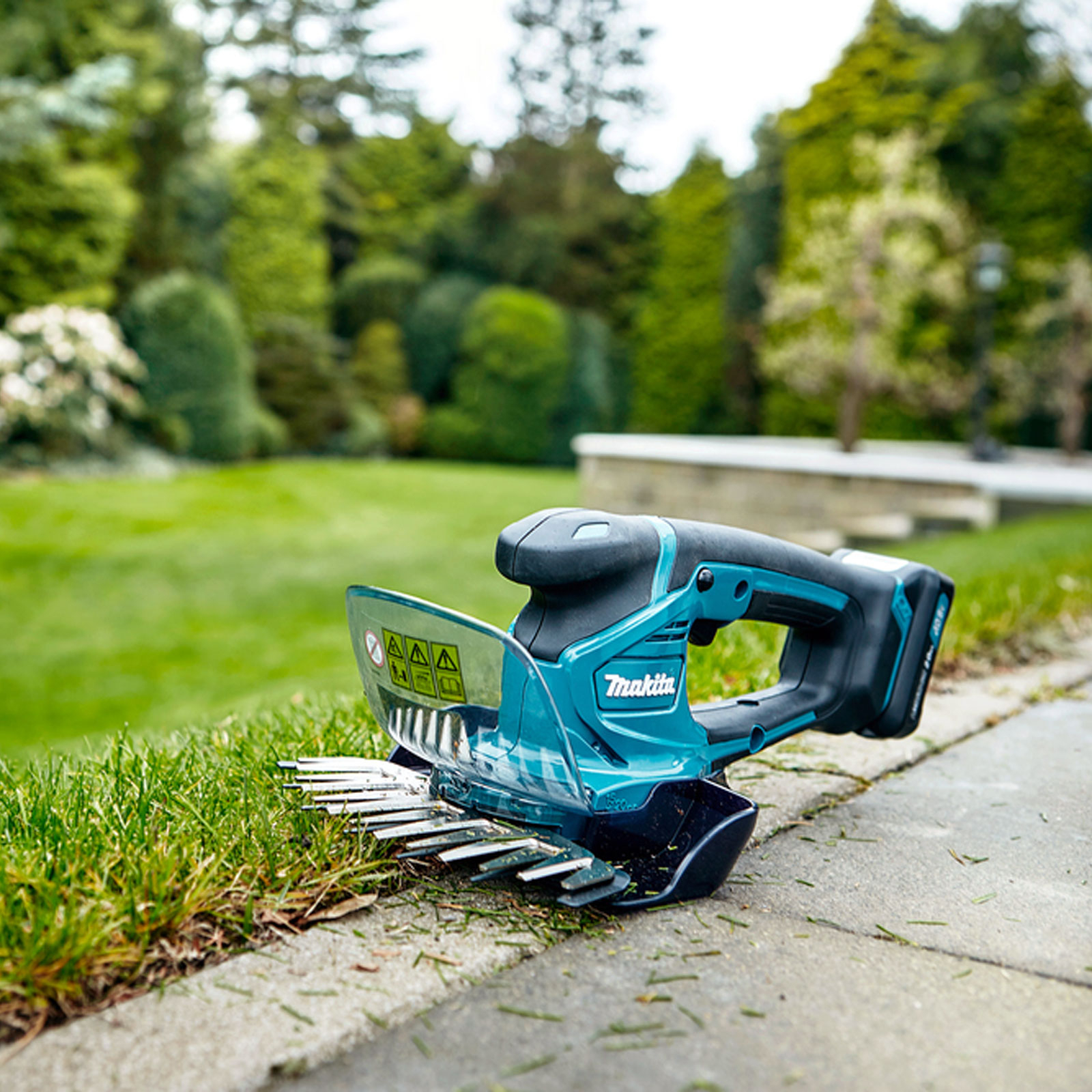 A teal and black Makita hedge trimmer rests on a concrete path next to green grass, with trees in the background.