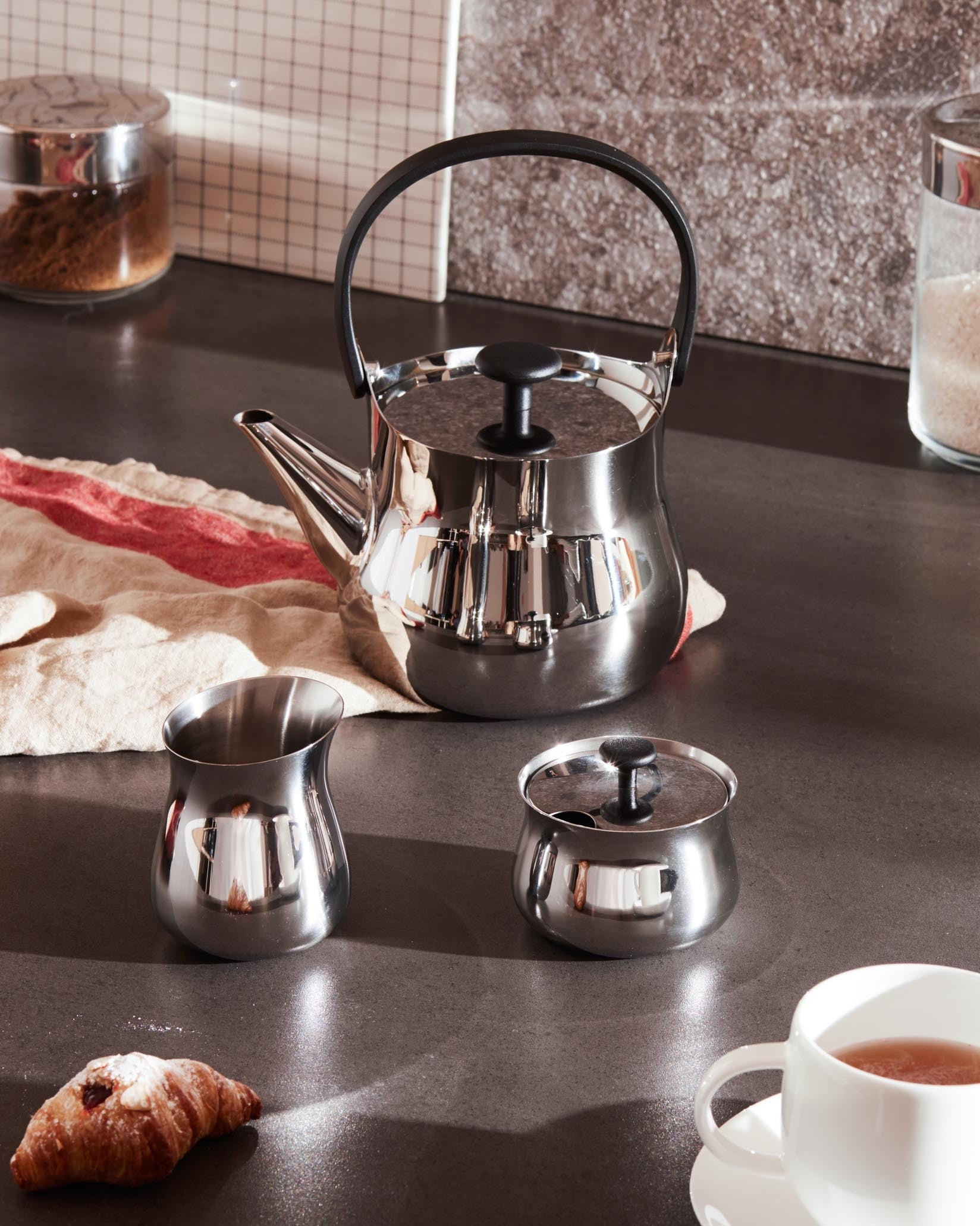 Un set da tè lucido include teiera, panna, zuccheriera e una tazza di tè con croissant. Lo sfondo ha barattoli.