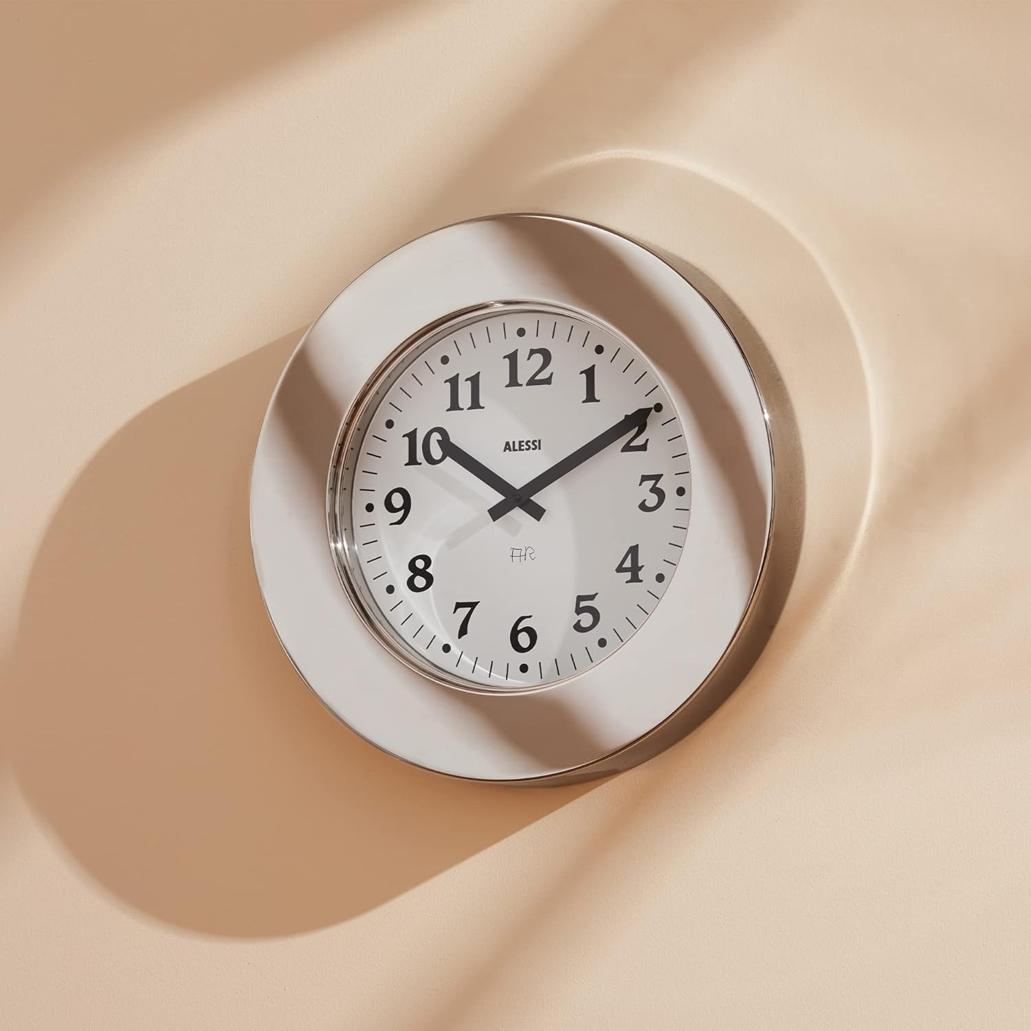 Un orologio rotondo con quadrante bianco e numeri neri è appeso a una parete beige. L'orologio è di Alessi.