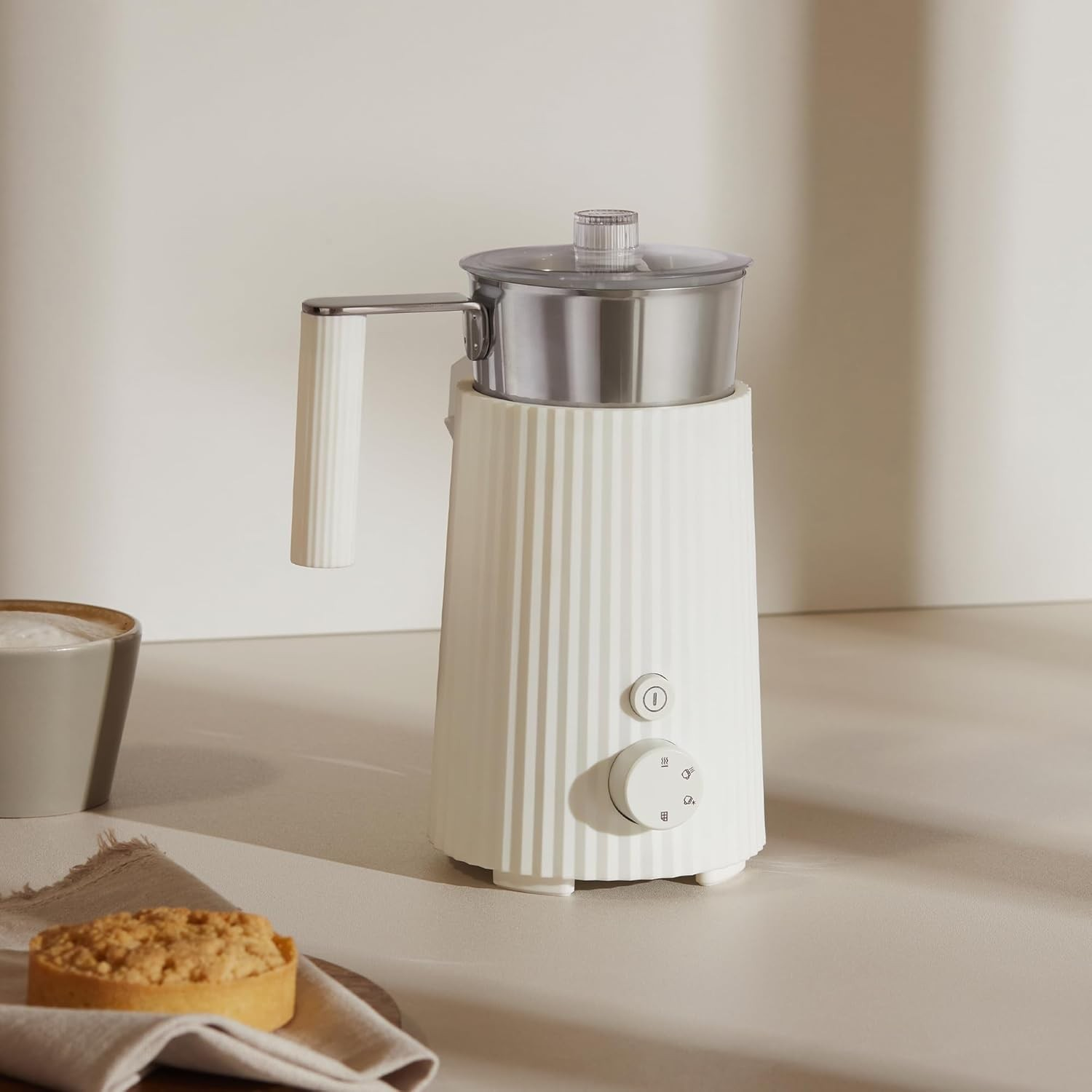 Un montalatte bianco con parte superiore, manico e comandi in metallo. Una tazza di caffè e una crostata sul tavolo.