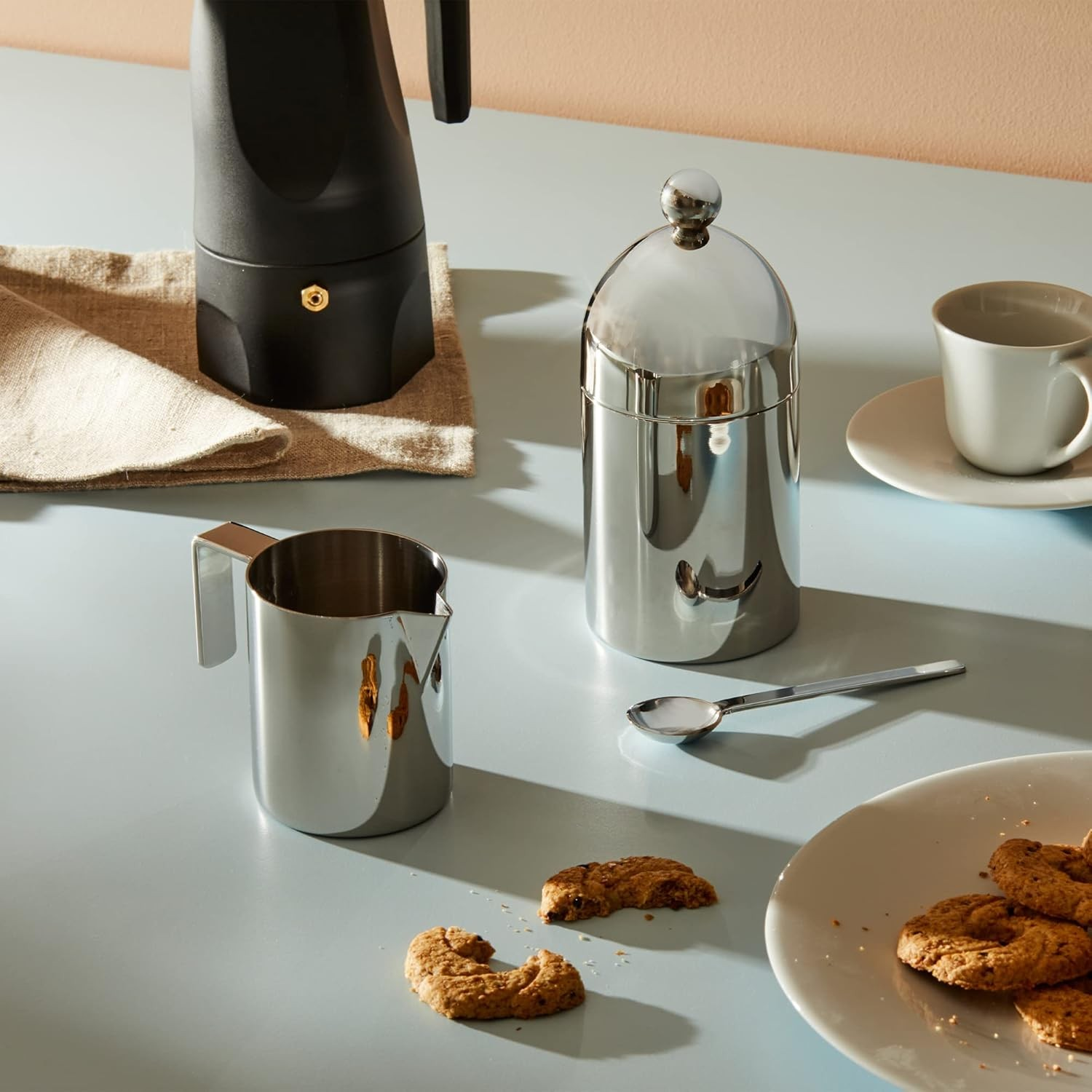 Una tavola apparecchiata con caffettiera, panna, zuccheriera, biscotti, tazza e cucchiaio. Lo sfondo è azzurro e pesca.