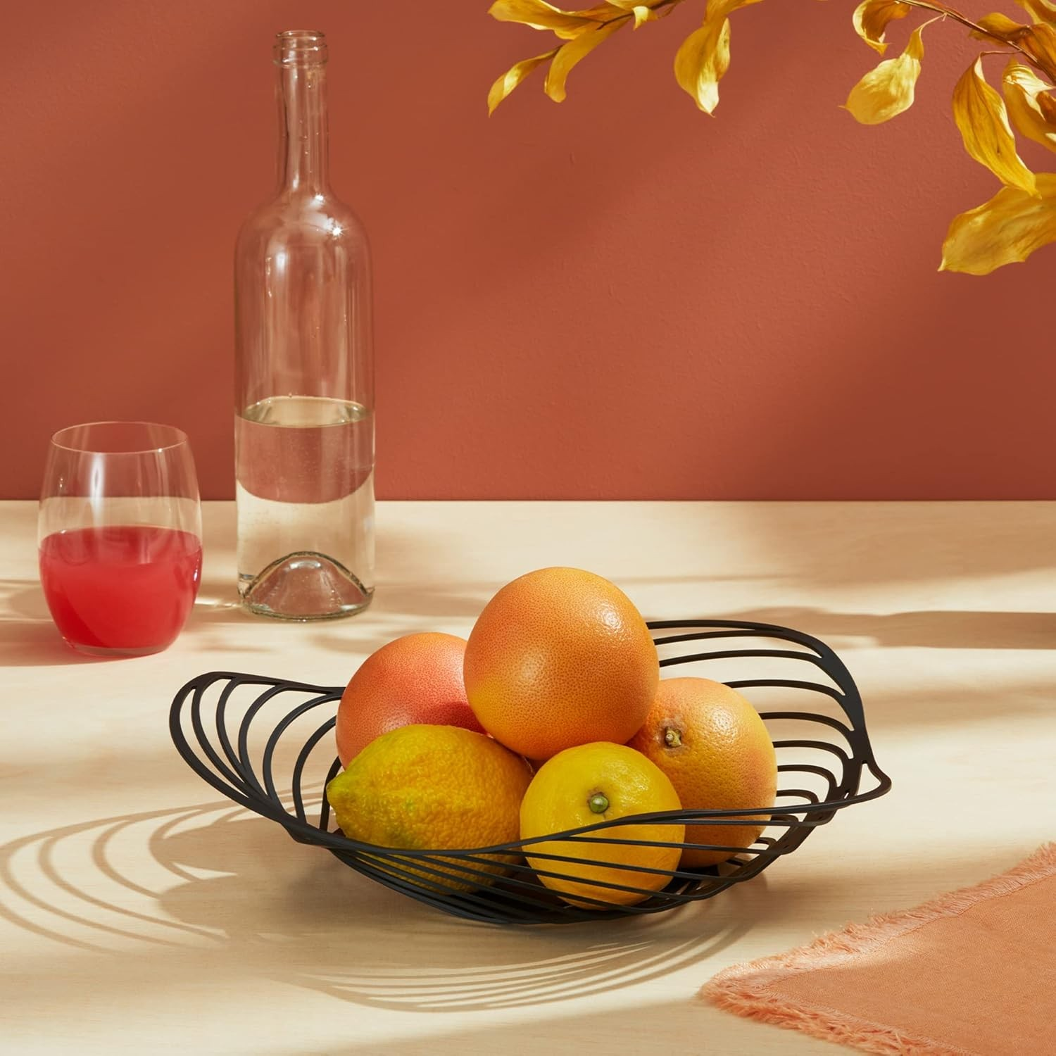 Un cestino di frutta nero contiene arance e limoni. Una bottiglia d'acqua e un bicchiere di succo sono su un tavolo. Lo sfondo è terracotta.