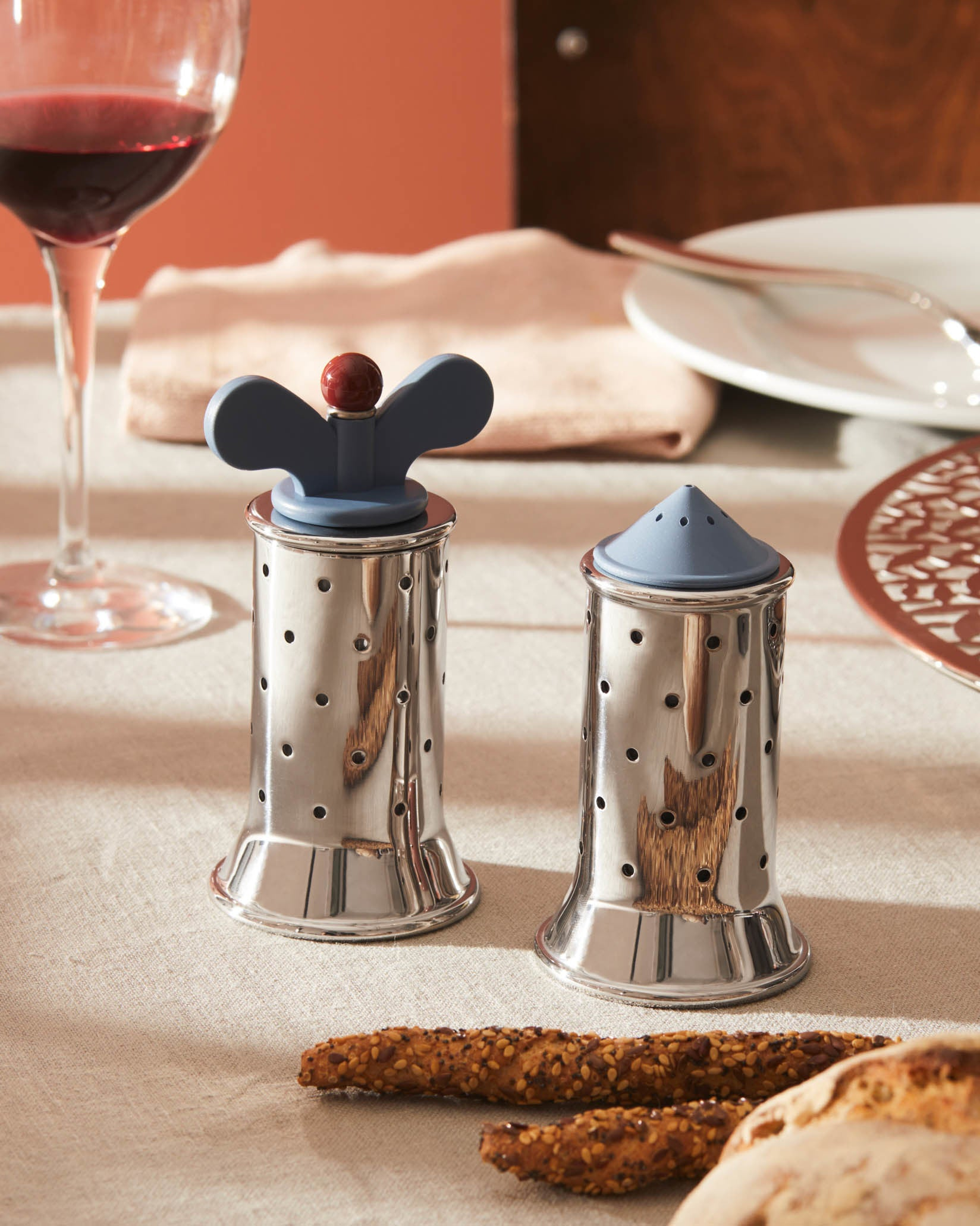 Due shaker di metallo su un tavolo. Grissini e pane in primo piano. Un bicchiere di vino e un piatto sullo sfondo.