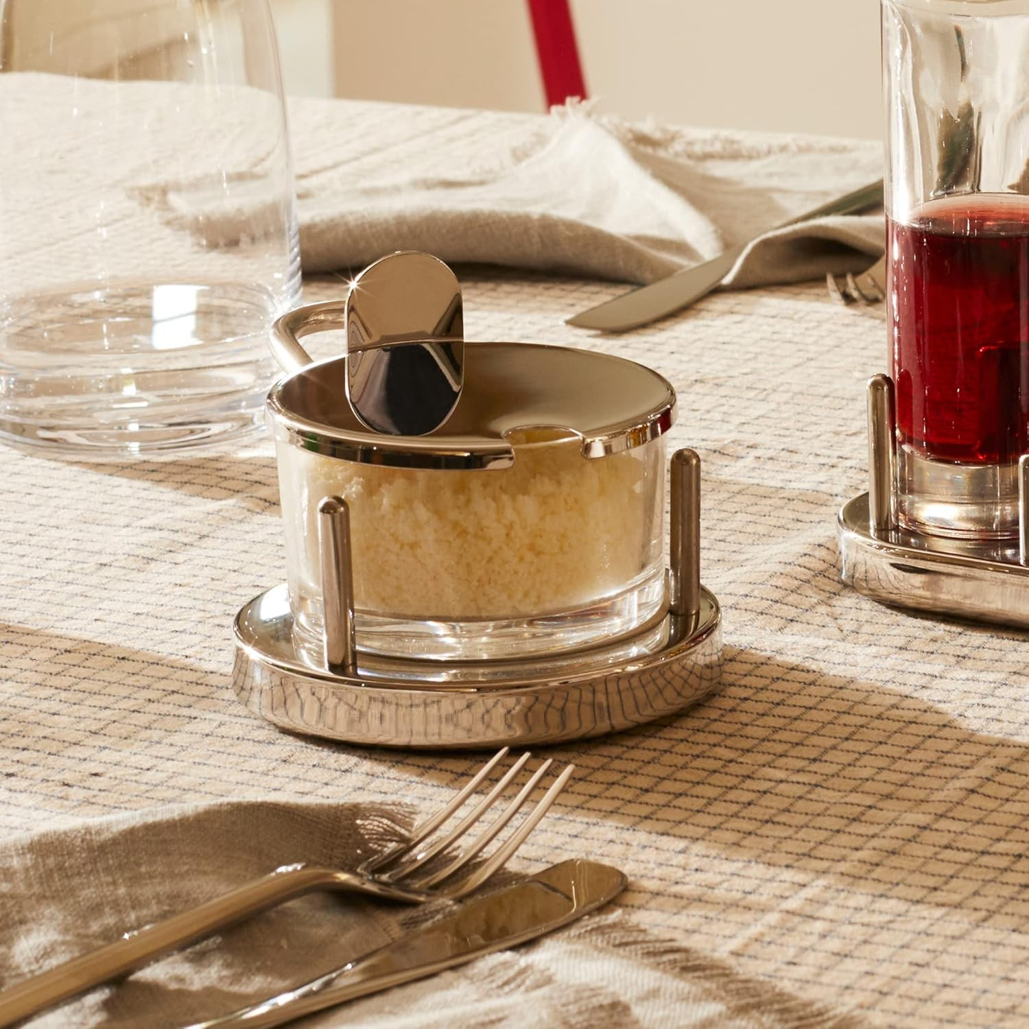 Una tavola apparecchiata con un contenitore per formaggio in vetro, un bicchiere di liquido rosso e posate su una tovaglia fantasia.