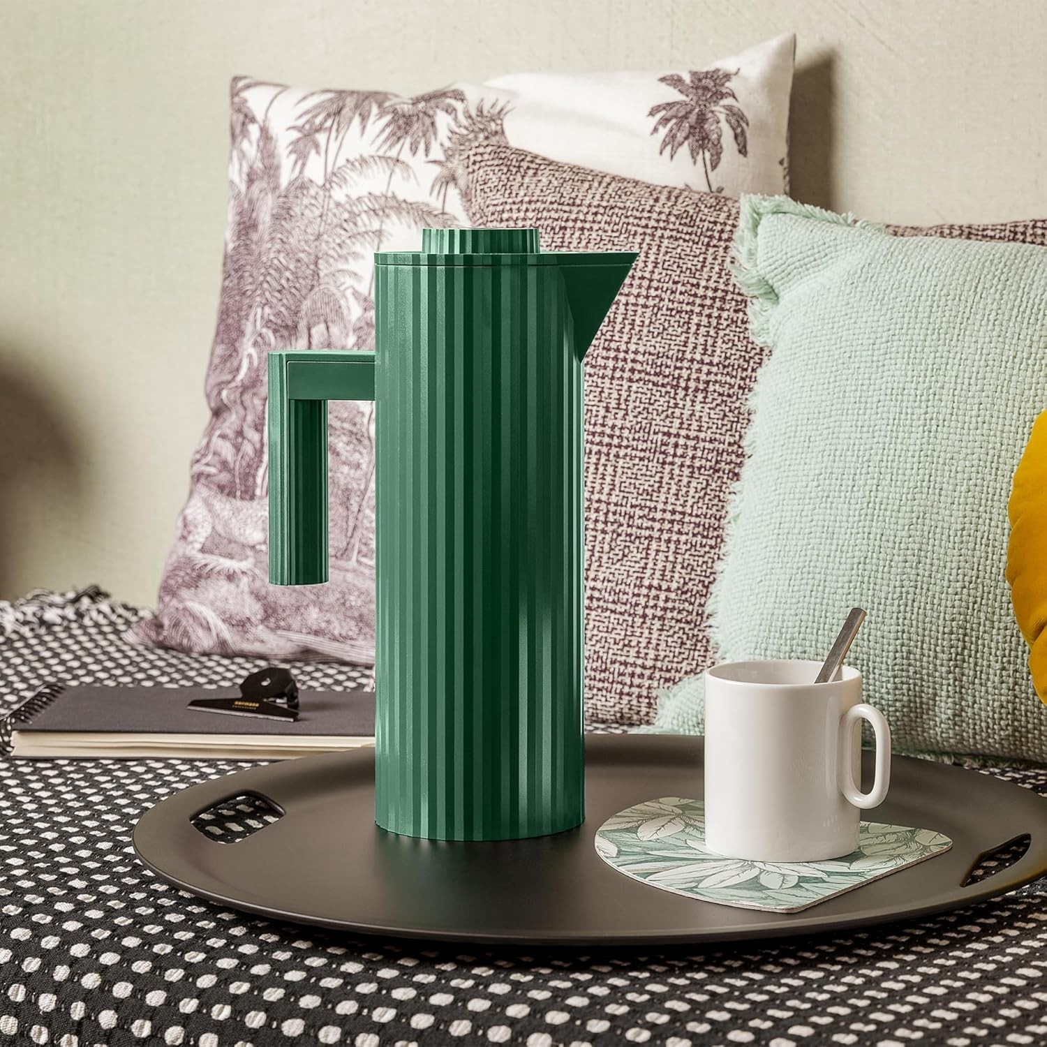 Una caffettiera verde e una tazza bianca con cucchiaino su un vassoio. Cuscini sullo sfondo.