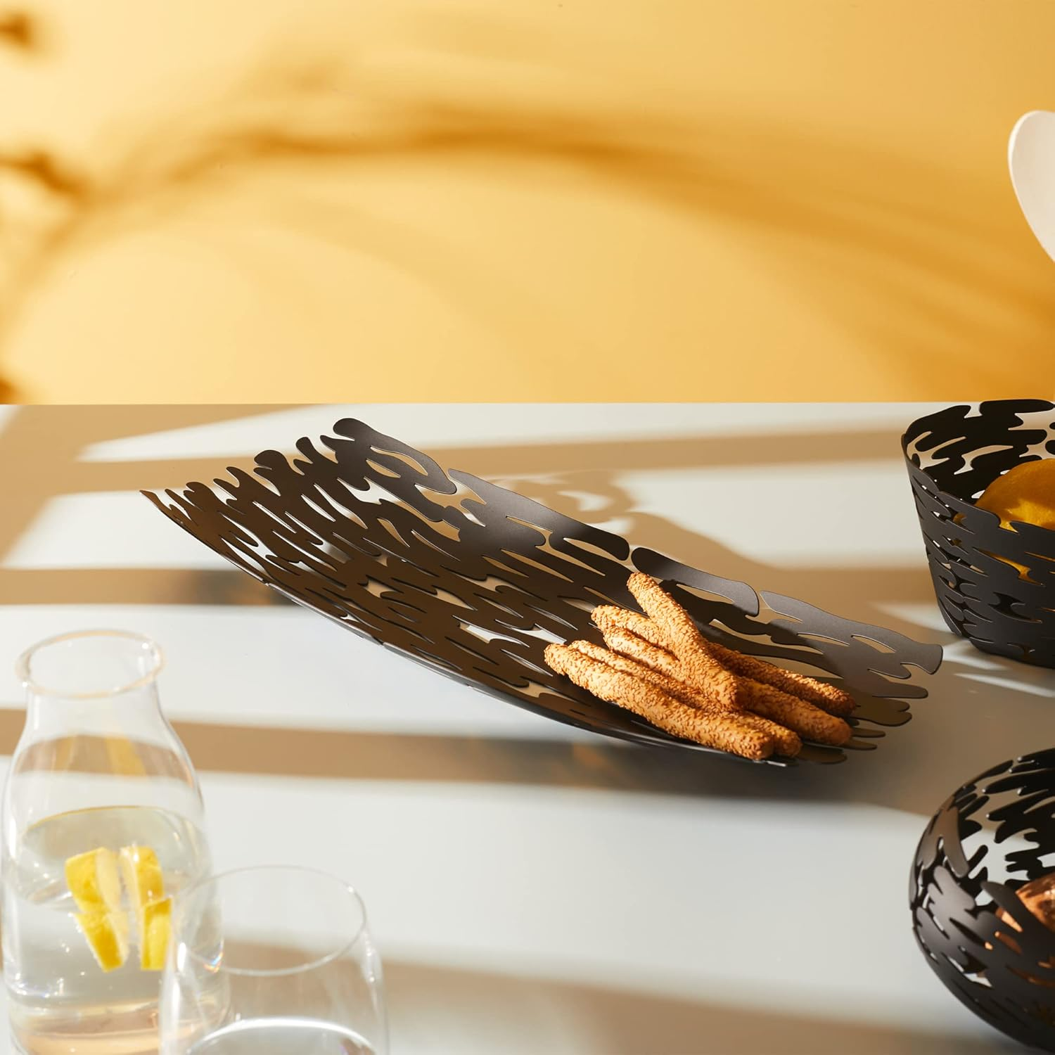 Un tavolo con un lungo vassoio decorativo con grissini, una piccola ciotola decorativa nera, una bottiglia e un bicchiere d'acqua con limone.