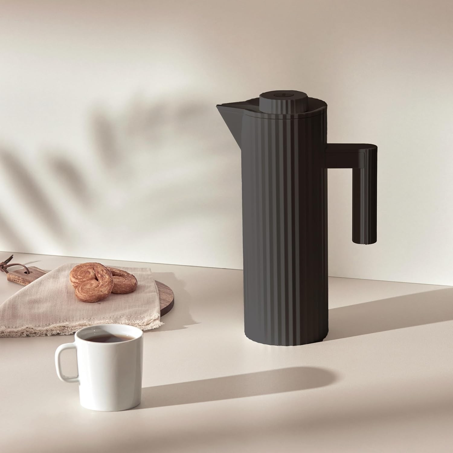 Una caffettiera francese nera, tazza bianca con caffè, biscotti e un tagliere in legno su una superficie bianca con ombre.