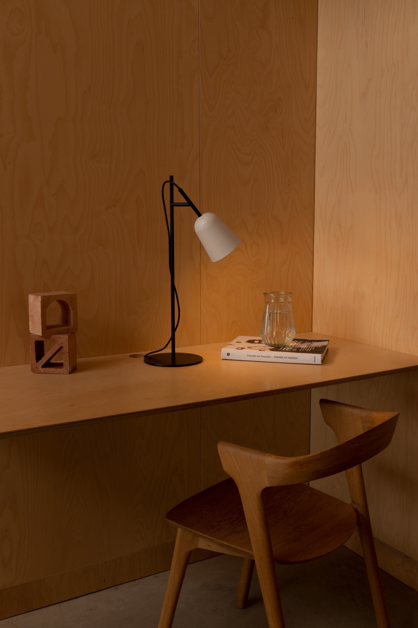 Una scrivania con una lampada bianca, un bicchiere d'acqua e un libro. C'è una sedia di legno e dei blocchi di legno sulla scrivania.