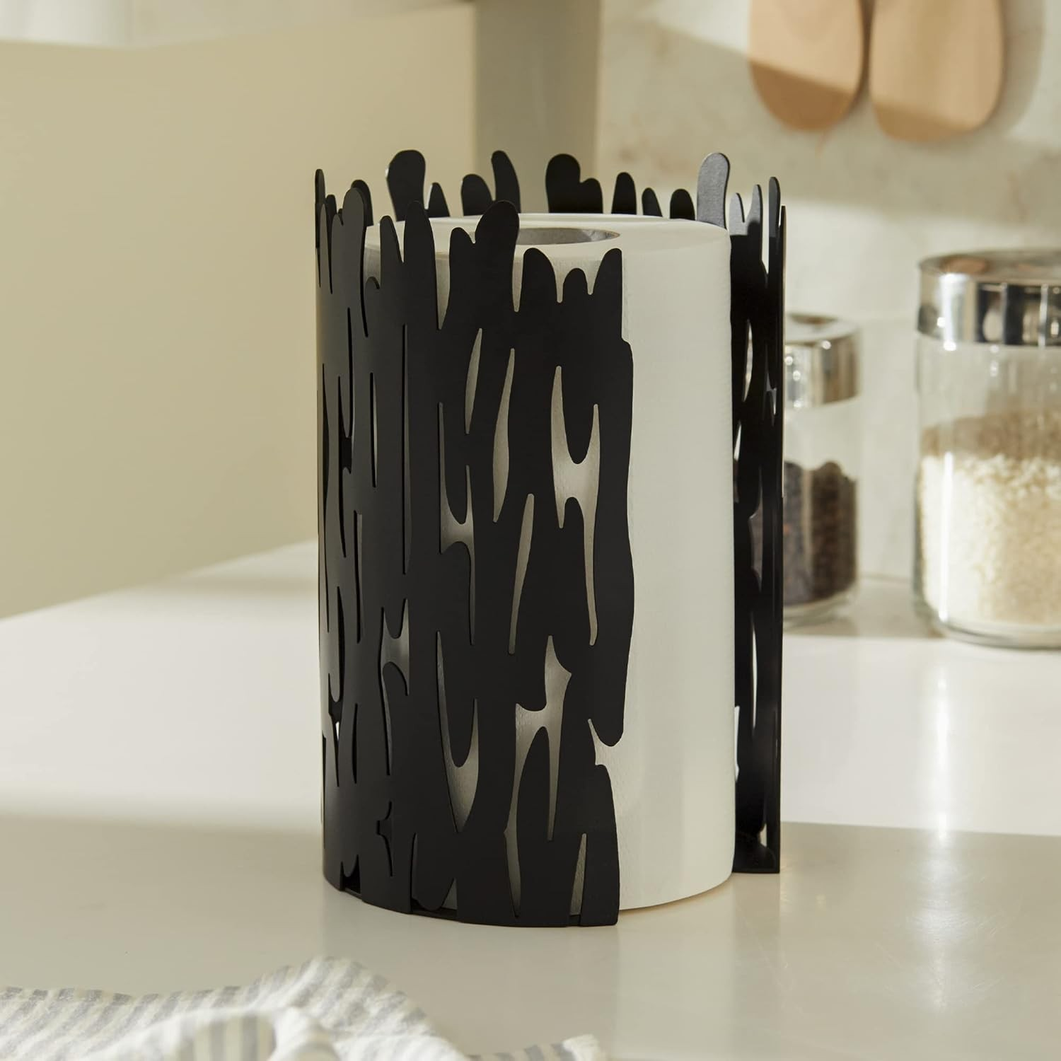 Un cilindro bianco è racchiuso da un manicotto decorativo in metallo nero. Lo sfondo presenta barattoli, cucchiai di legno e una superficie bianca.