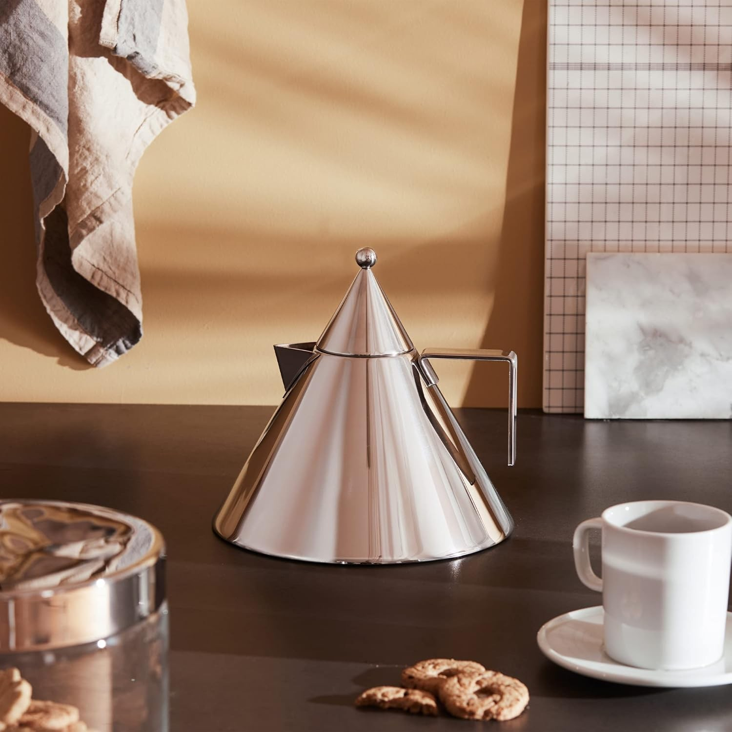 Una teiera conica lucida è appoggiata su una superficie scura accanto a una tazza bianca e biscotti. Sono presenti anche un barattolo di biscotti e un panno appeso.
