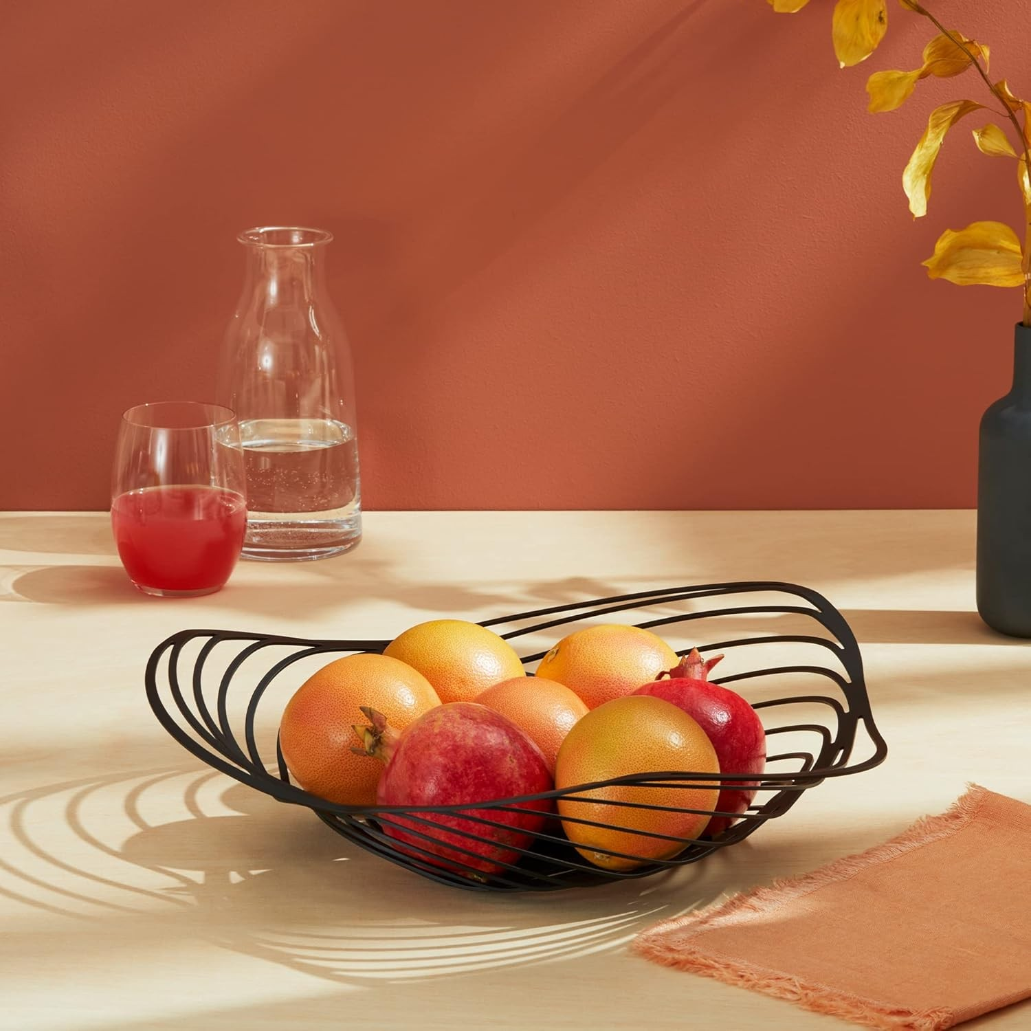 Una fruttiera in filo nero contiene pompelmi e melograni. Un bicchiere di succo, una caraffa d'acqua e un vaso con foglie gialle sono vicini.