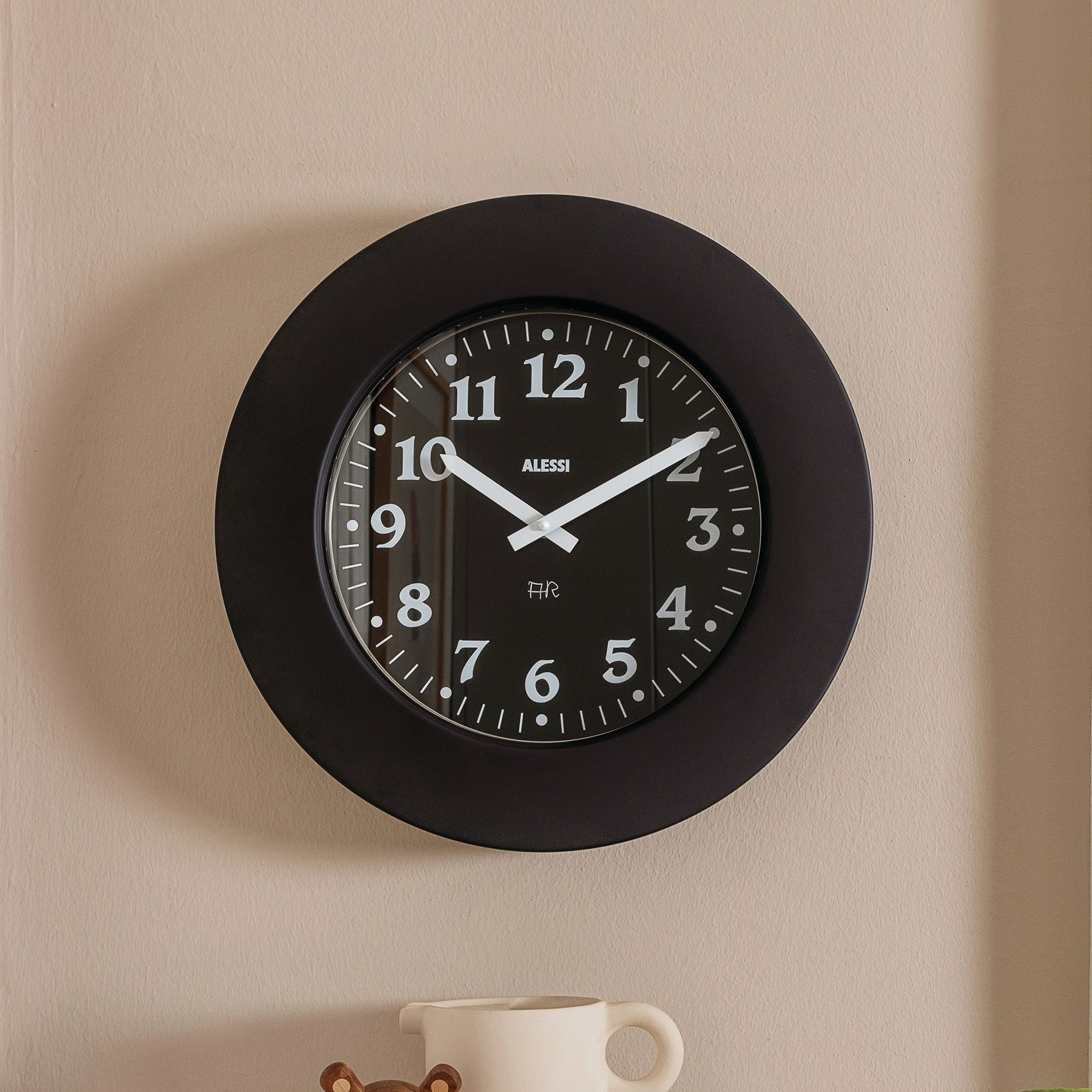 Un orologio Alessi nero su un muro beige segna le 10:08. Una tazza bianca e un giocattolo di legno sono in primo piano.
