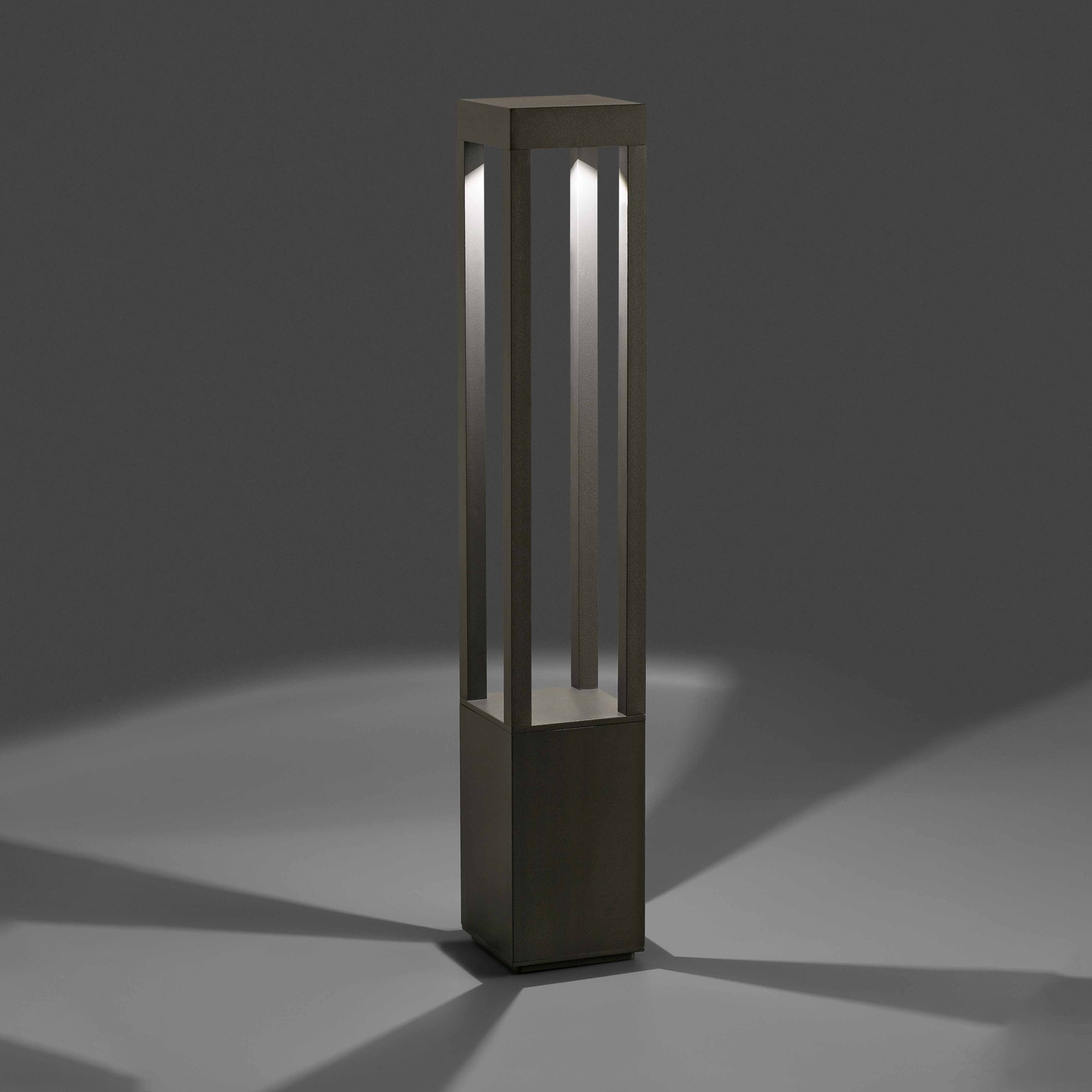 Una moderna lampada da esterno grigio scuro. È rettangolare con barre verticali, illuminata dall'interno. Le ombre sono proiettate sul pavimento.